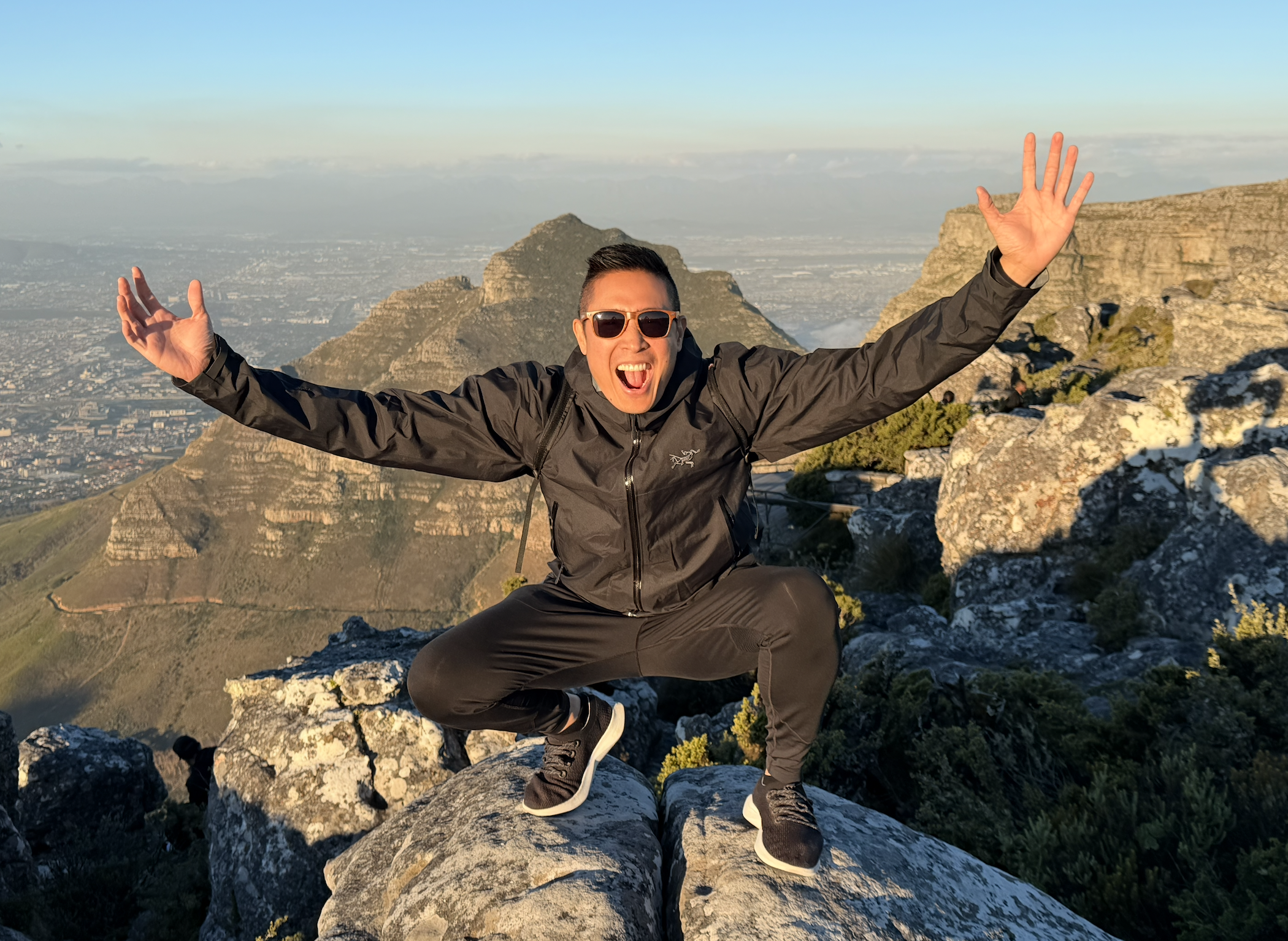 George Luc wearing sunglasses and a dark jacket is on a rocky mountain summit (table mountain), with mountains and a cityscape in the background, raising his arms in celebration.
