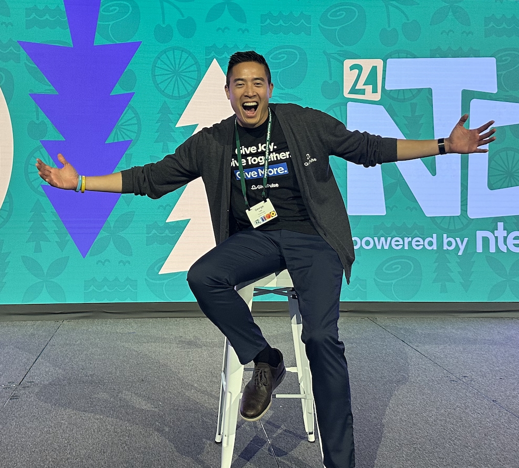 George Luc sitting on a stool on a stage with arms wide open, smiling enthusiastically, in front of a colorful background of the Nonprofit Technology Conference.