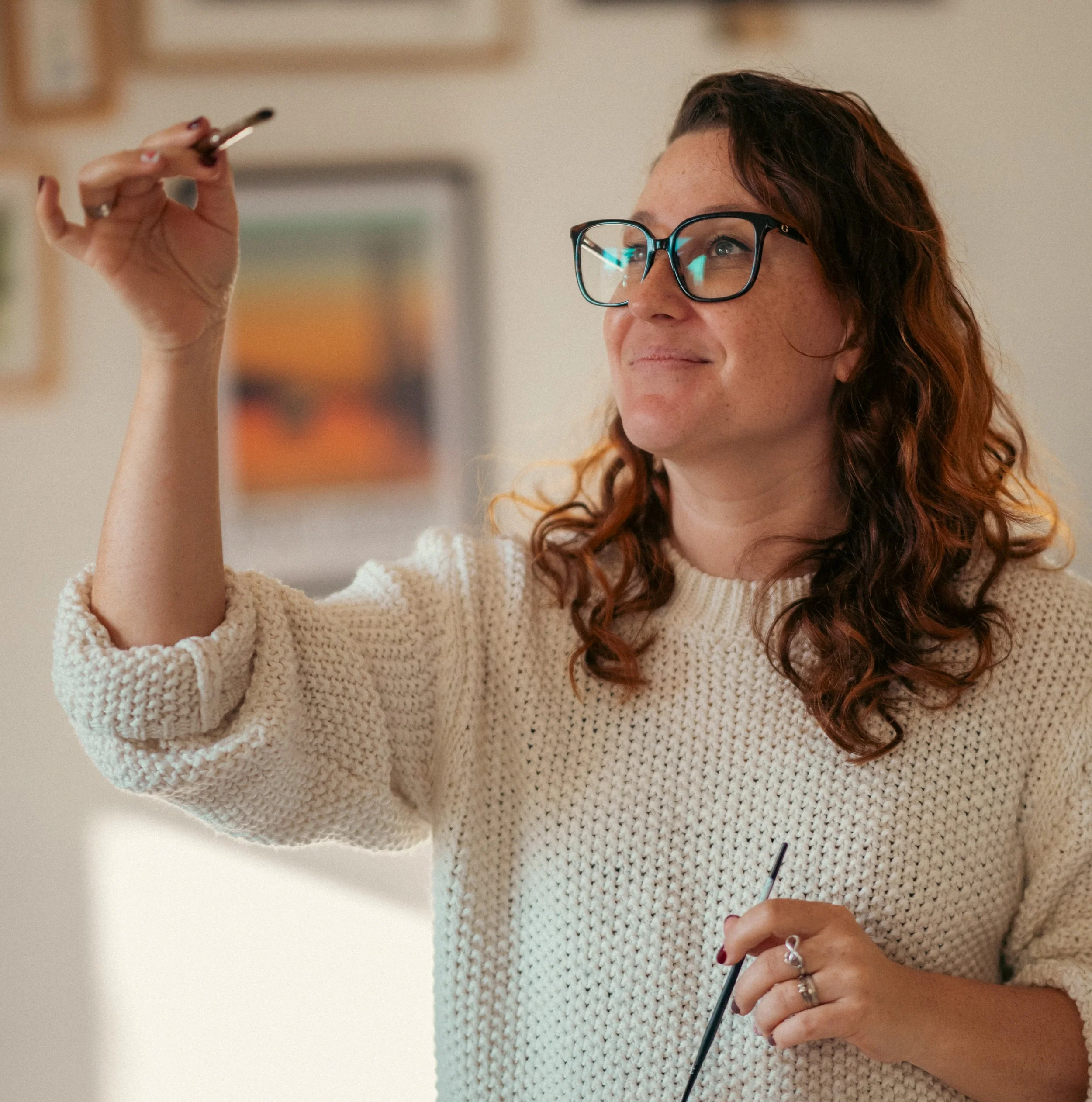 Une femme aux cheveux bouclés roux, portant des lunettes et un pull beige, tient un pinceau en main et le levant, dans une salle d'exposition ou un studio, avec des œuvres d'art en arrière-plan.