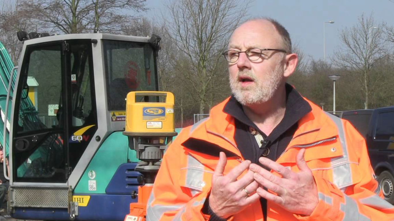 Engineer in an orange high-visibility jacket standing in front of excavation equipment, reviewing InSAR ground motion data for the site