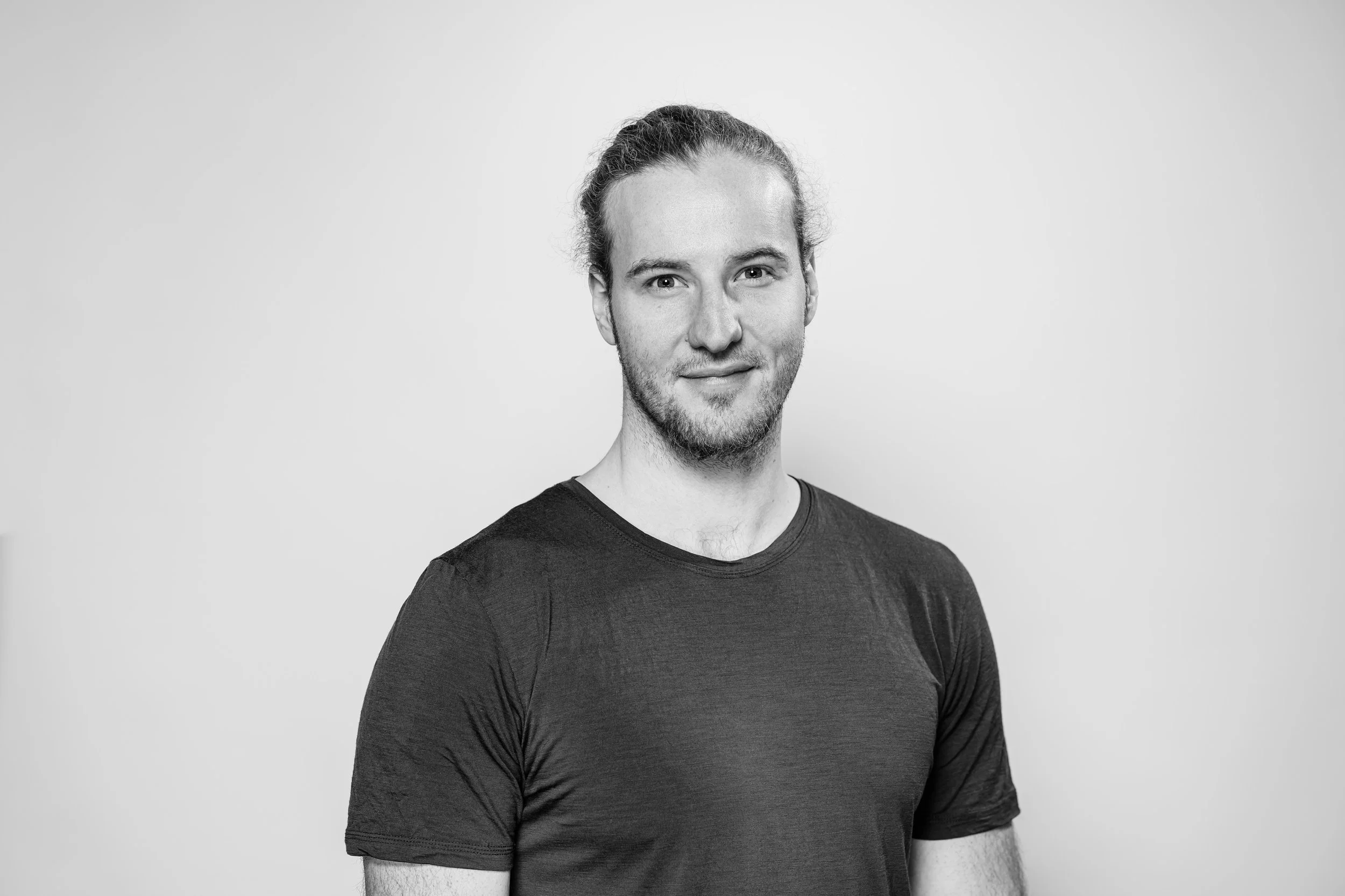 Black and white portrait of a young man with long hair tied back, wearing a plain dark T-shirt, standing against a plain light background.