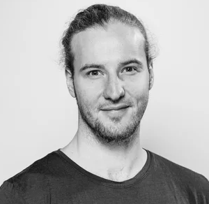 Black and white portrait of a young man with long hair tied back, wearing a plain dark T-shirt, standing against a plain light background.