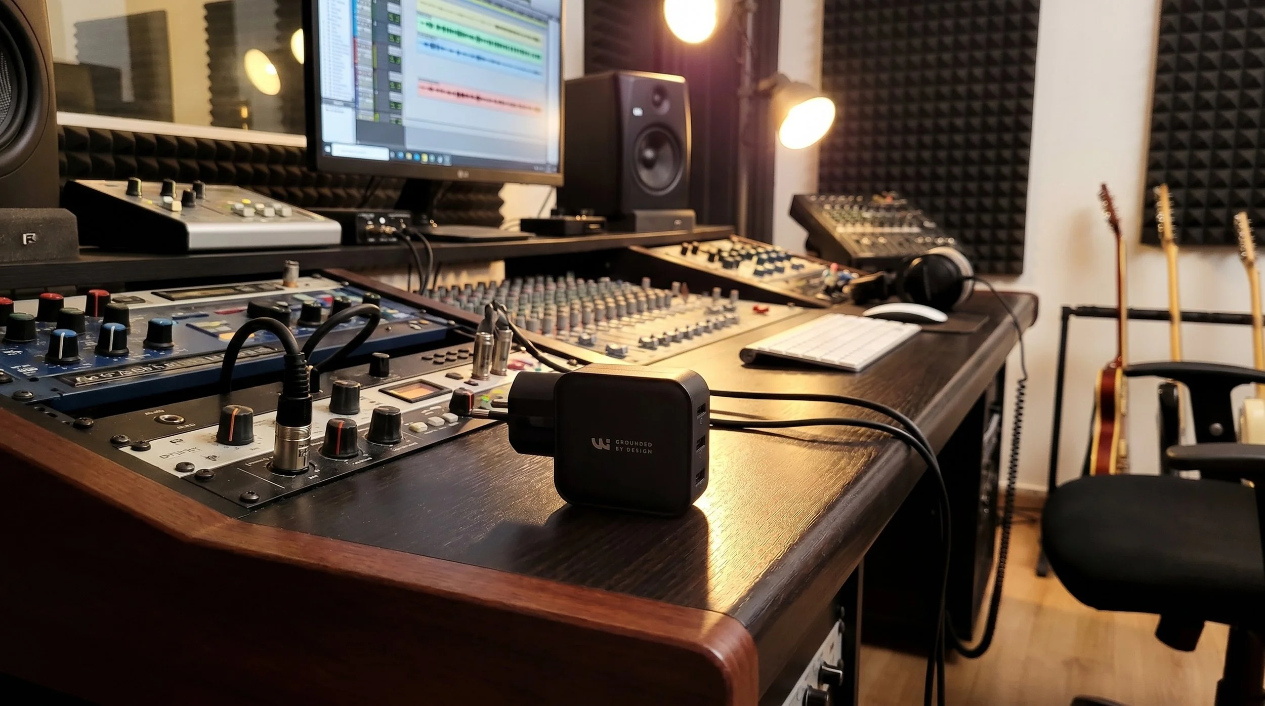 Studio d'enregistrement avec des équipements audio, des haut-parleurs, un ordinateur avec une station de travail numérique, des guitares en arrière-plan, des écouteurs, et une table de mixage. Chargeur wicaya sur la table