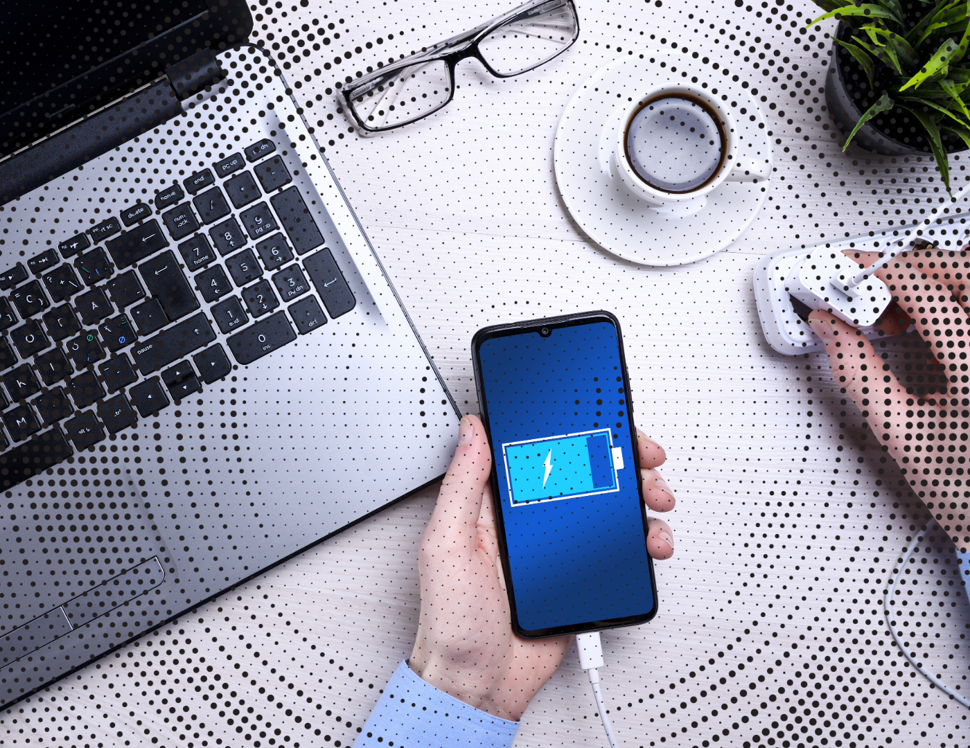 Une personne recharge un smartphone avec une batterie faible sur un bureau blanc avec un ordinateur portable, des lunettes, une tasse de café, une plante et une main utilisant un appareil laser.