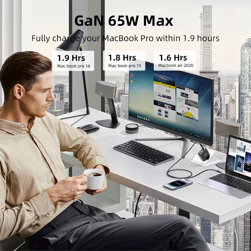 Un homme assis à un bureau avec un ordinateur, un téléphone, et une tasse de café, devant une grande fenêtre avec vue sur la ville. Le texte indique la durée de charge d'un MacBook Pro à 65W Max.