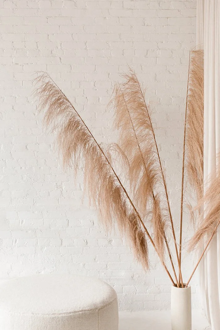 Beige pampas grass arrangement in a white vase next to a white wall with a textured fabric on the right side.