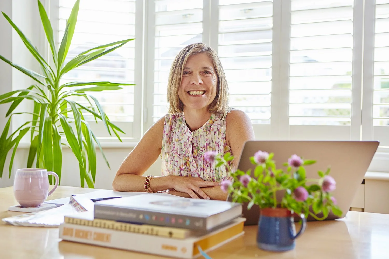 UK Squarespace website designer, Nora Rose Zinerman, sitting at a table, smiling, with a laptop, design books and a tea, in front of a window full of light.