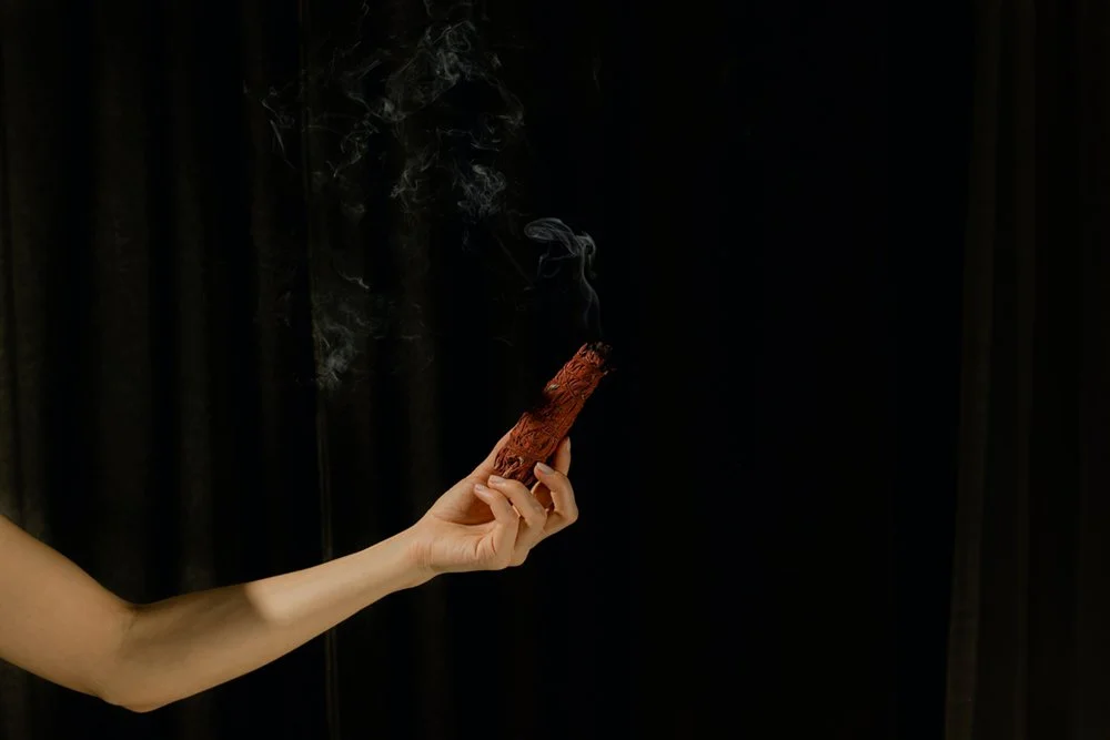 A person's hand holding burning sage smudge stick with smoke rising against a dark background.