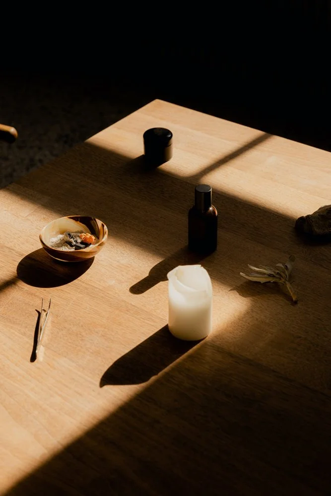A wooden table with a white candle, a small dark bottle, a bowl with an assortment of herbs or incense, a pair of tongs, and a single flower. Sunlight creates shadows on the table.