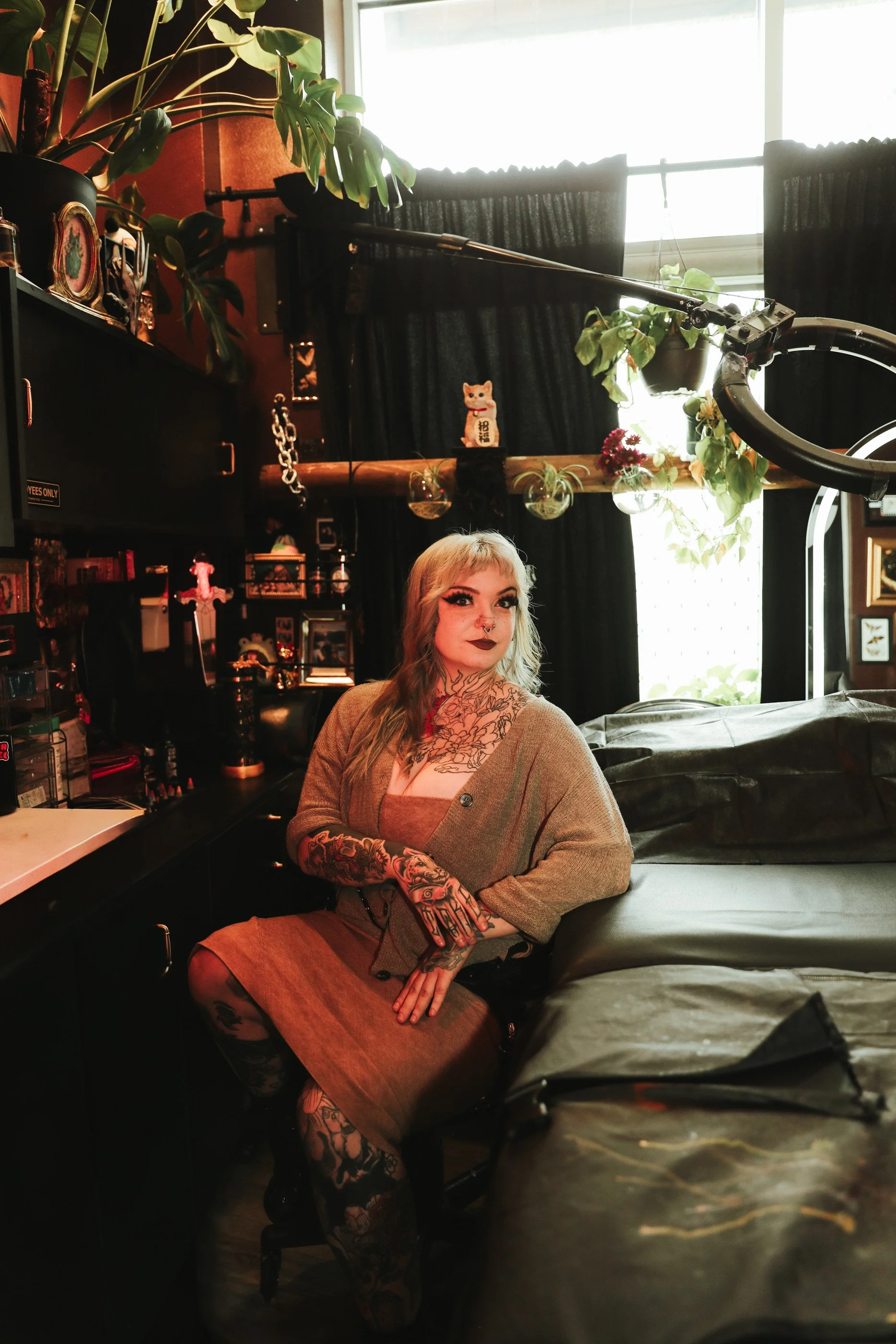 A woman with light skin, blonde and brown hair, and multiple facial pierces, including a septum and nostrils, is smiling at the camera. She is wearing a headband with decorative crystals and makeup with dark eyeliner. Next to her is a dog with a brown face, black and white fur, and a big nose, wearing a patterned collar, and resting its head on her shoulder. The background shows a blue sky with some clouds and a part of a sign with red and green colors.