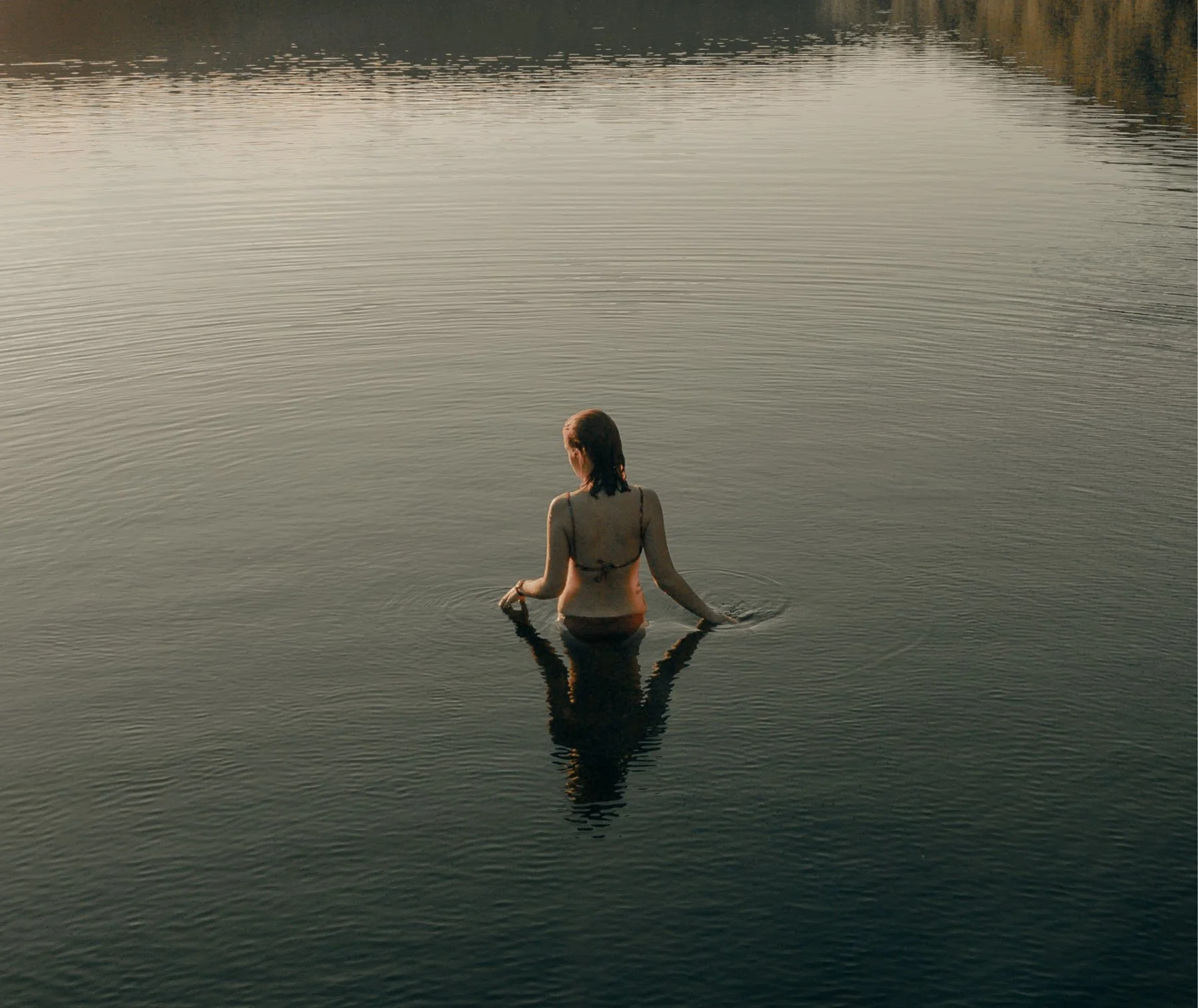 A woman standing waist-deep in calm lake water with her back facing the camera, during sunset or dusk, with trees in the distant background.