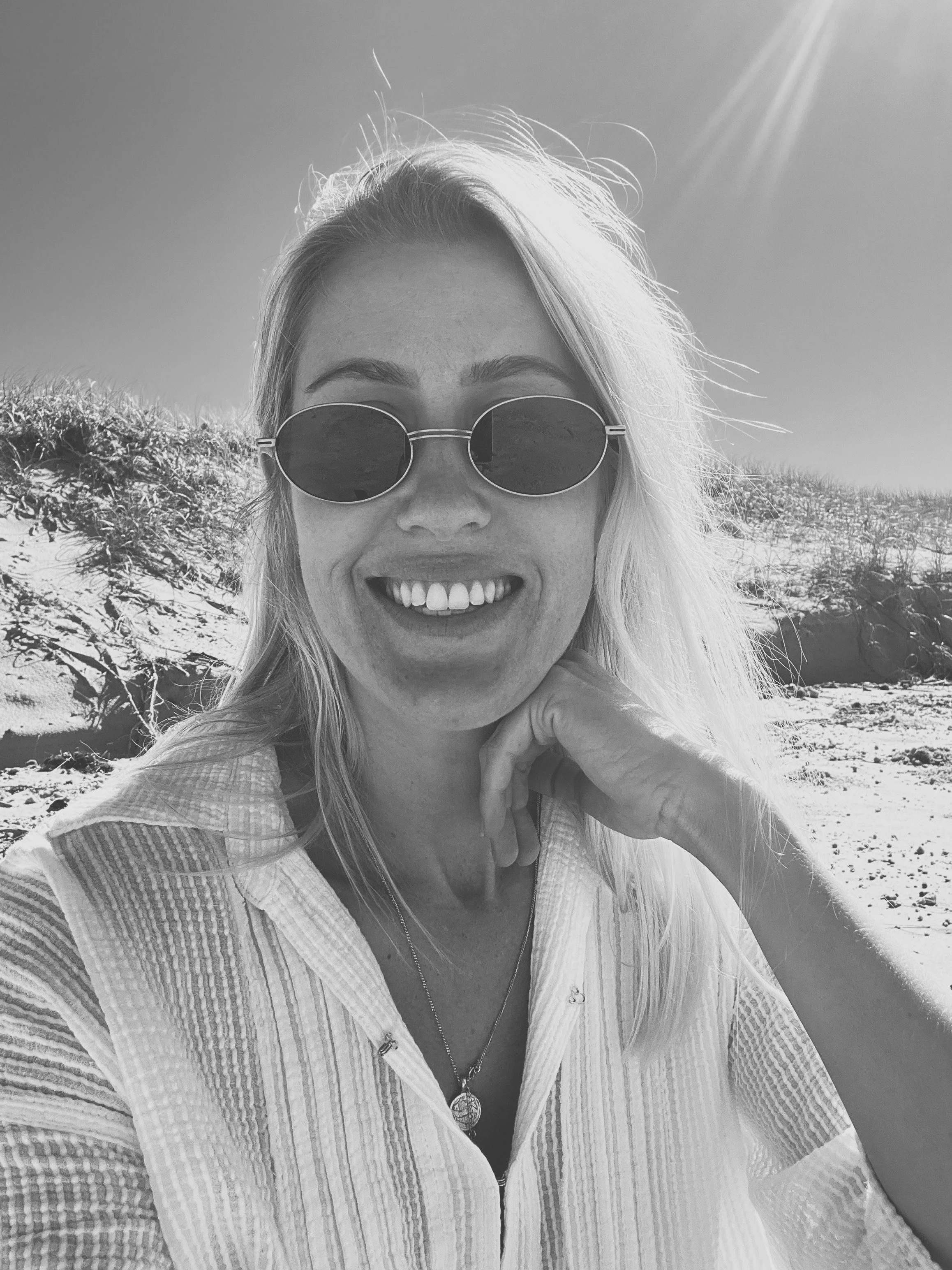 Black and white photo of the business founder with sunglasses and a necklace, outdoors on a sunny day with sand dunes in the background.
