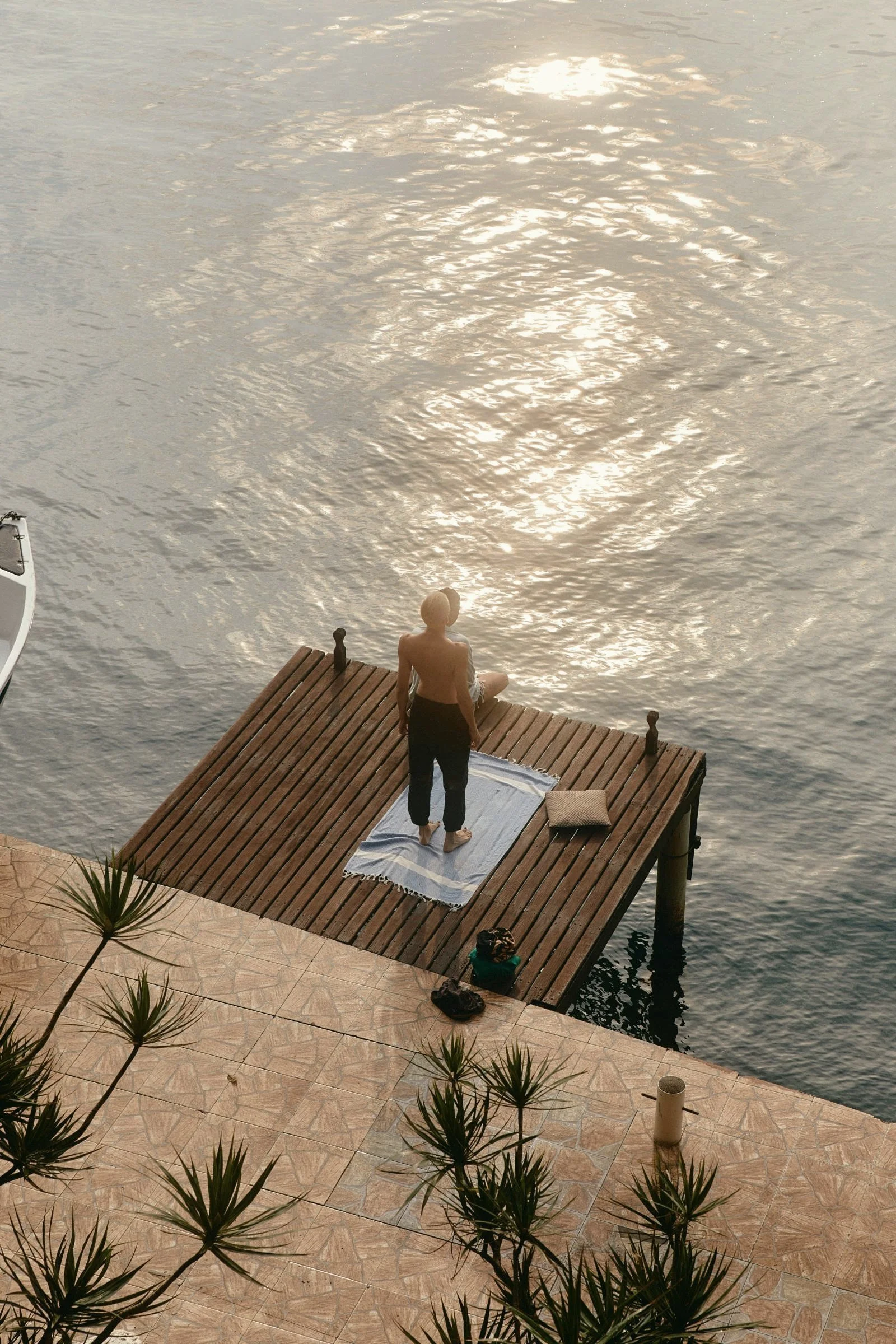 A person practicing yoga or meditation on a wooden dock by the water during sunset or sunrise, with a towel and pillow nearby, and plants in the foreground.