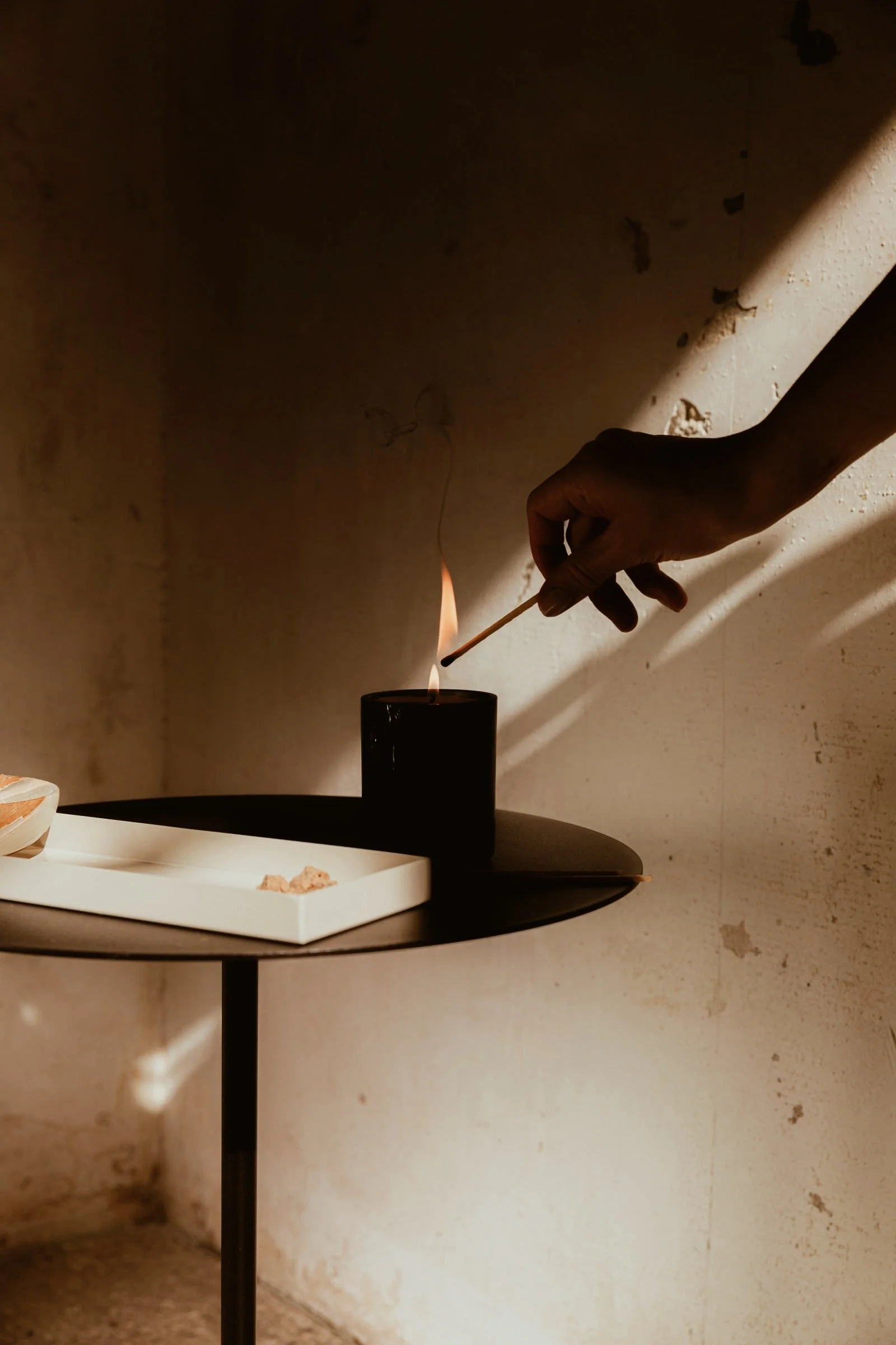 A person lighting a candle with a matchstick in a dimly lit room. There is a white tray on a black table in the foreground and an textured beige wall in the background.
