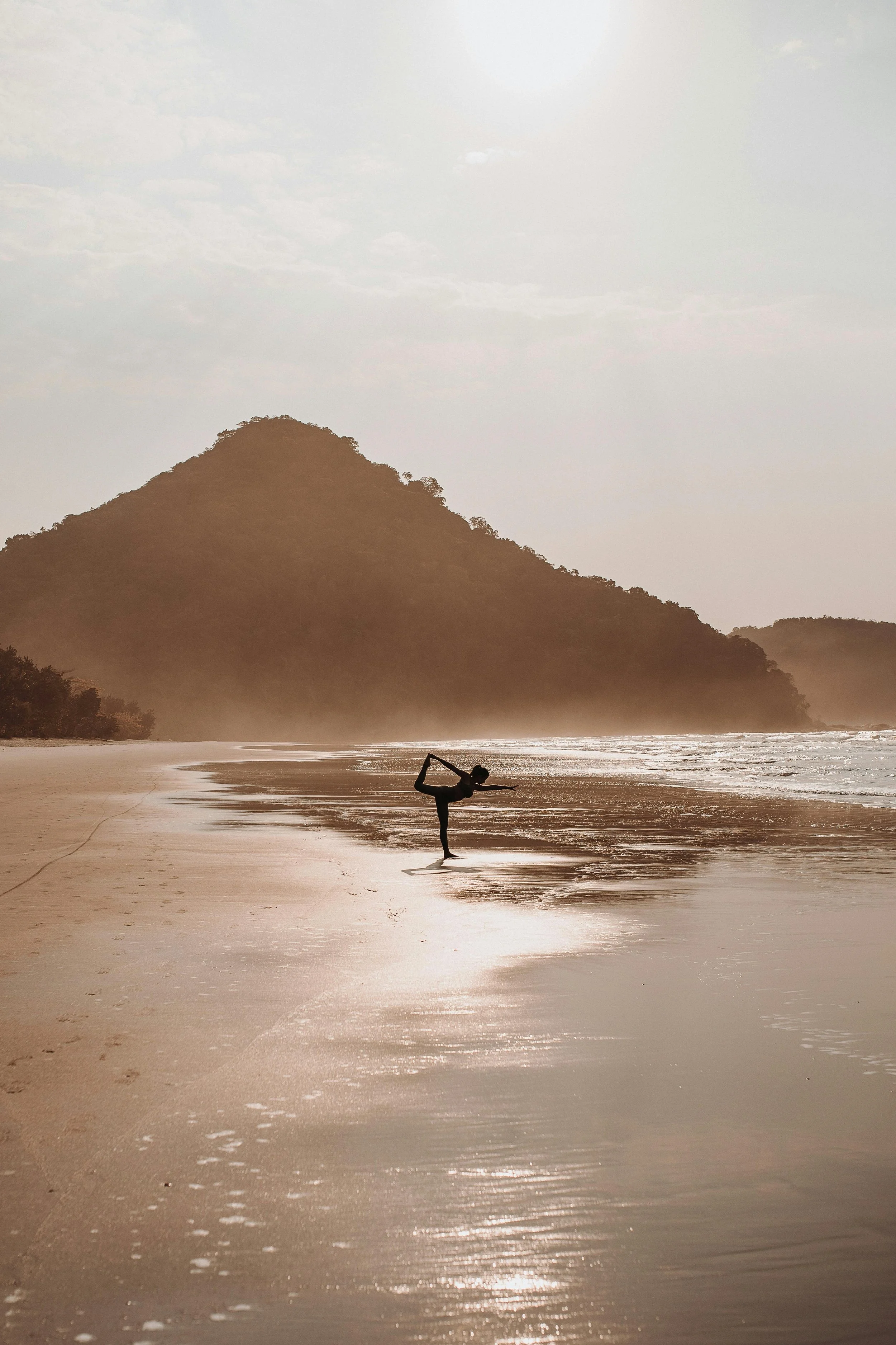 A person practicing yoga on a beach at sunset with mountains in the background.