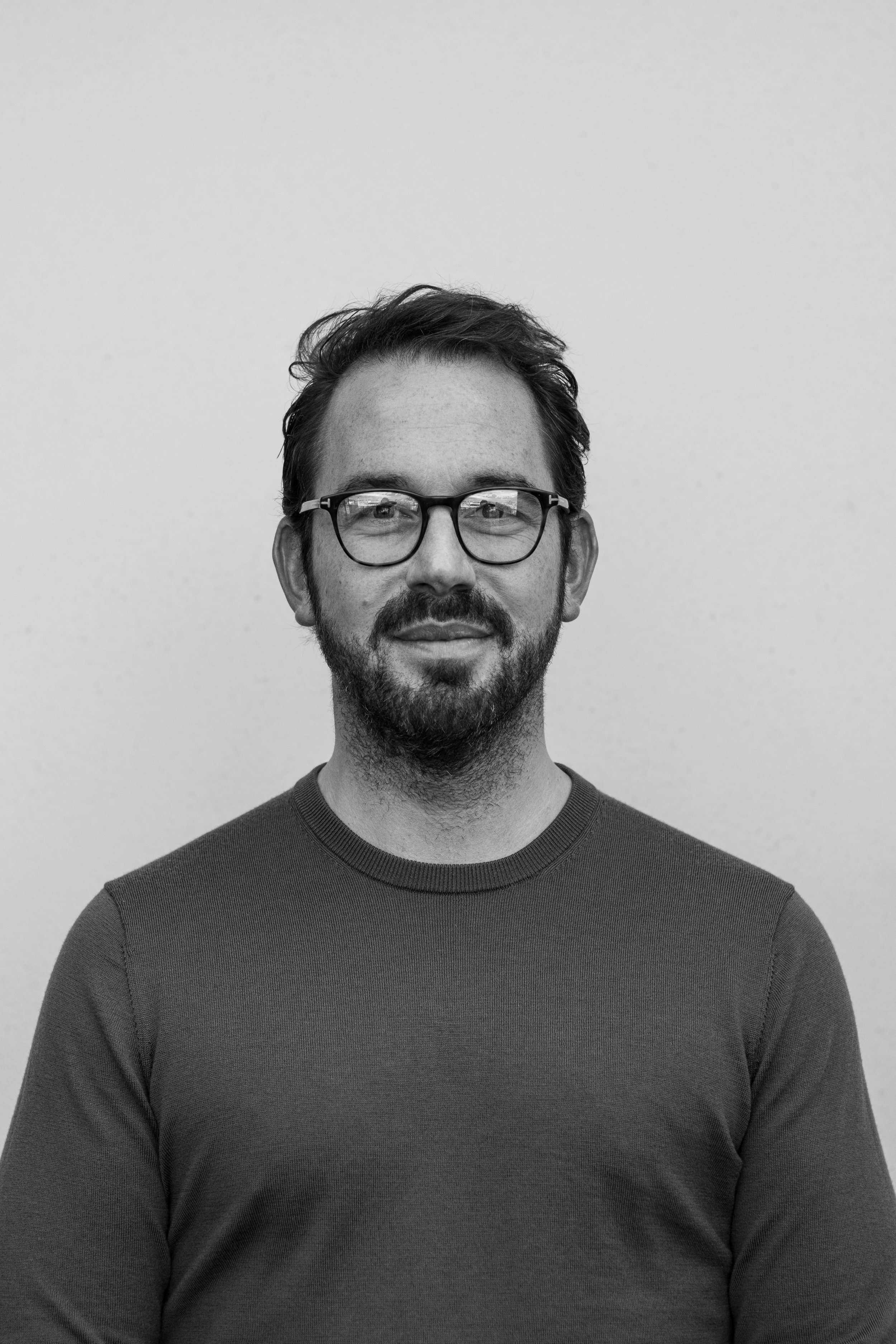 A black-and-white portrait of a man with glasses, a beard, and short hair, wearing a crew neck shirt, standing against a plain background.