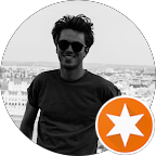 Homme souriant portant des lunettes, en extérieur avec une ville en arrière-plan, en noir et blanc.