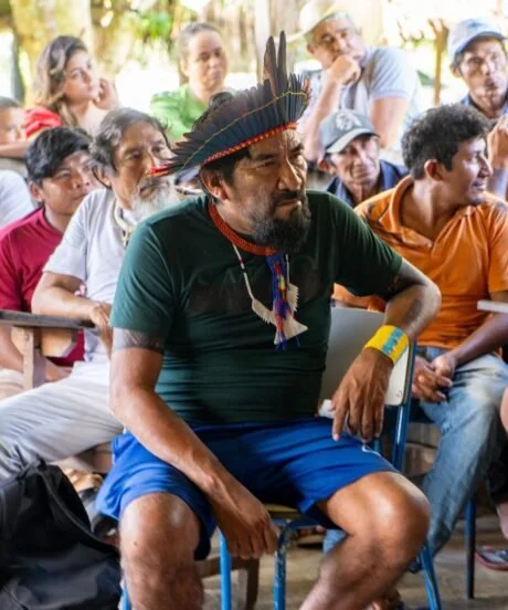 A man wearing a feathered headdress and traditional necklace sits in front of a diverse group of people, some with hats, in an outdoor setting.