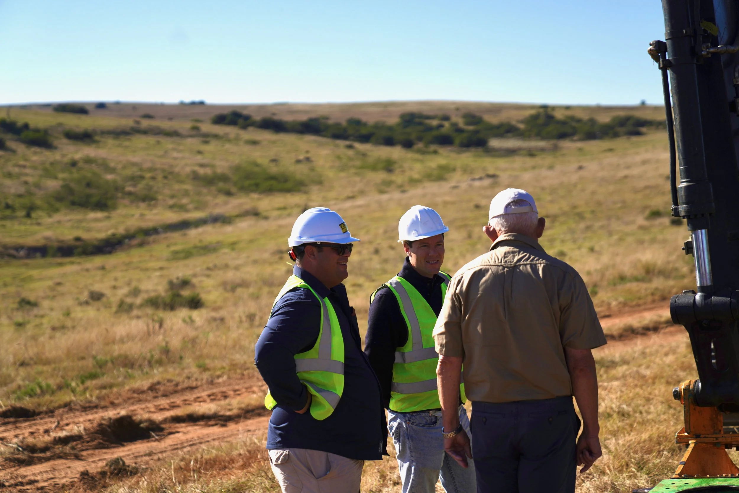 Three men wearing safety helmets and high-visibility vests talking outdoors on a grassy field with a clear sky.