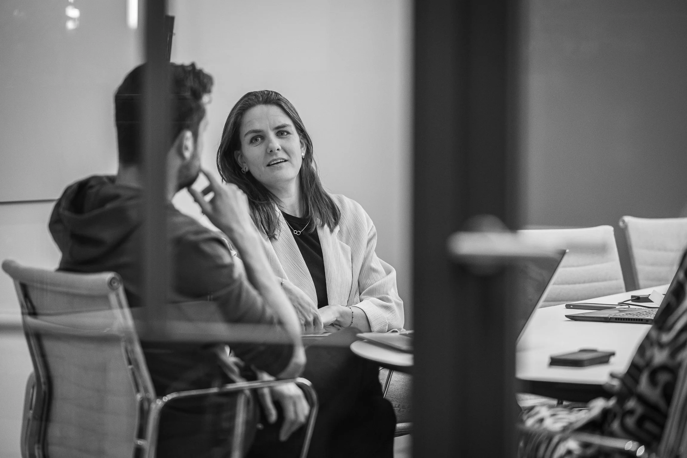 Black and white photo of two people in a meeting room, one woman facing the camera and talking, the other person with a beard listening, seated at a table with laptops and papers.