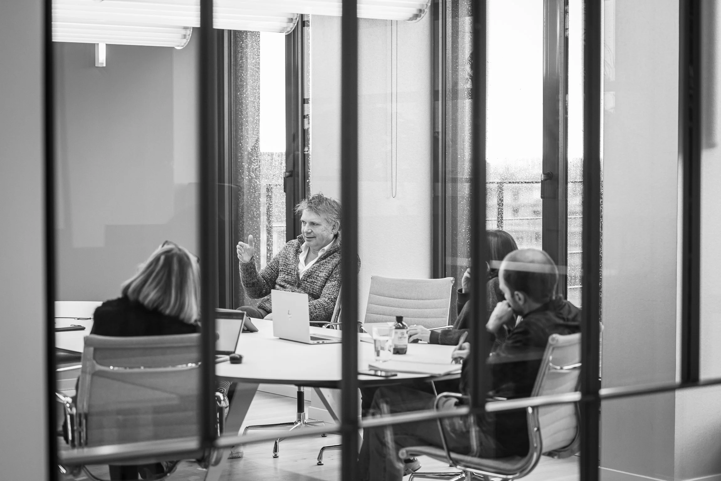 A black and white photo of four people having a meeting in a conference room, seen through glass walls. The person in the center appears to be speaking, while others listen.