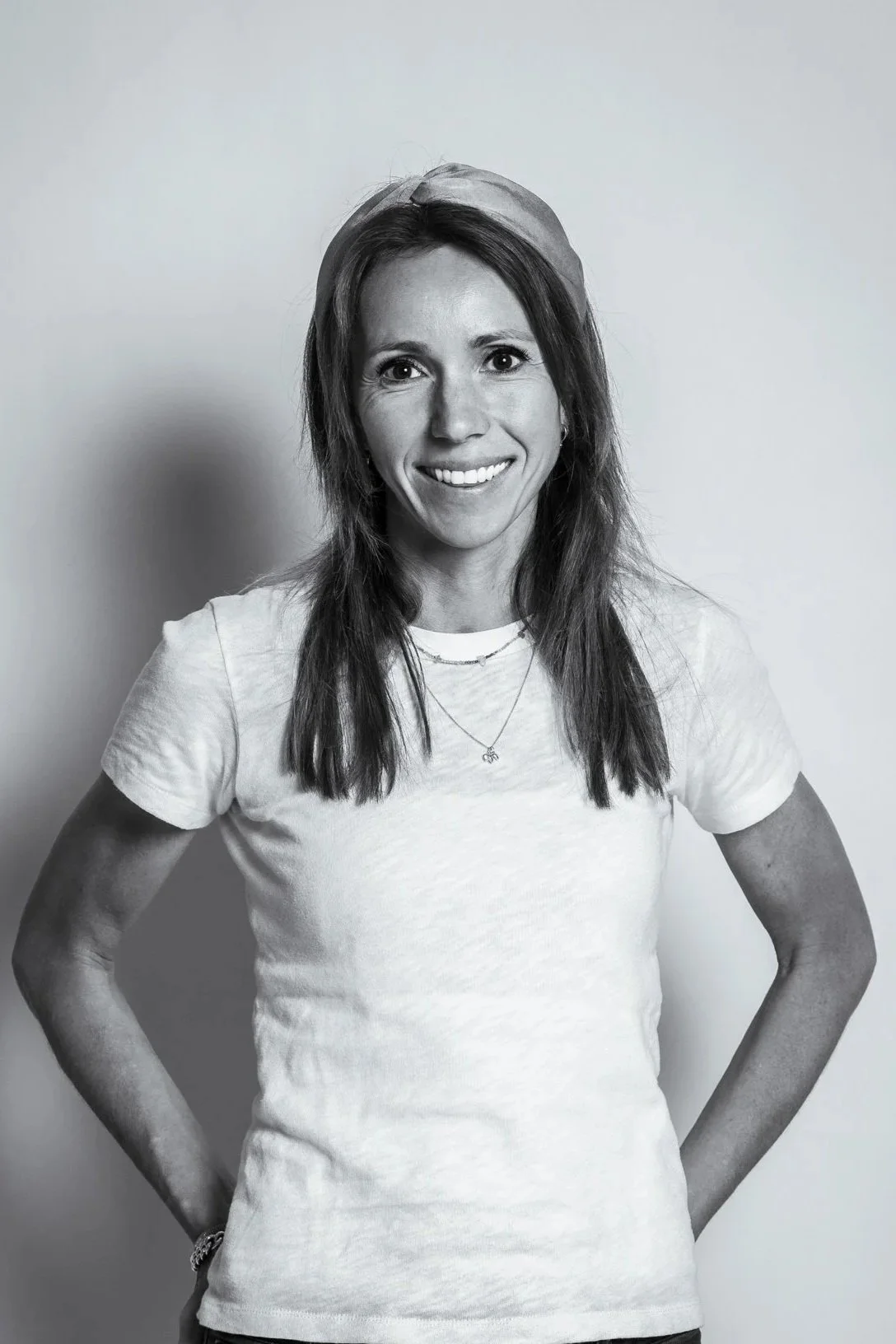 Black and white photo of a woman smiling with her hands on her hips, wearing a white t-shirt and layered necklaces, standing against a plain background.