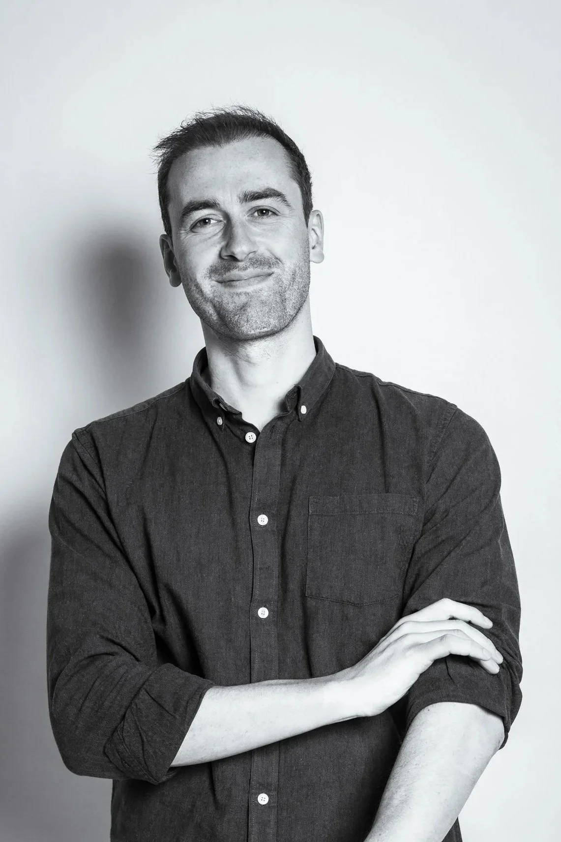 A black-and-white portrait of a man with short hair, slight beard, wearing a button-up shirt, smiling and crossing his arms in front of a plain wall.