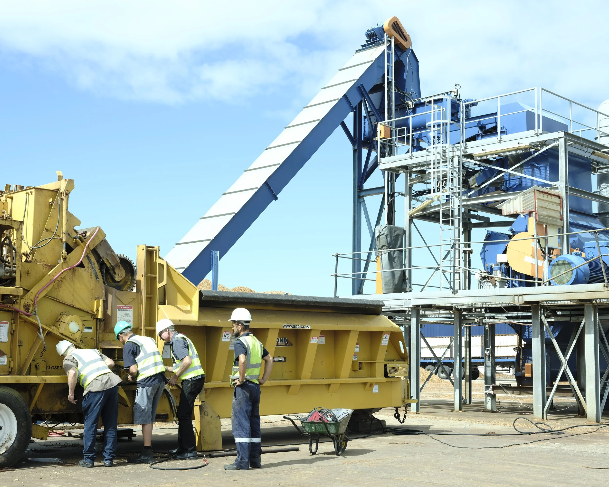 Four workers with safety helmets and vests operating machinery on a construction site with large industrial equipment and scaffolding.