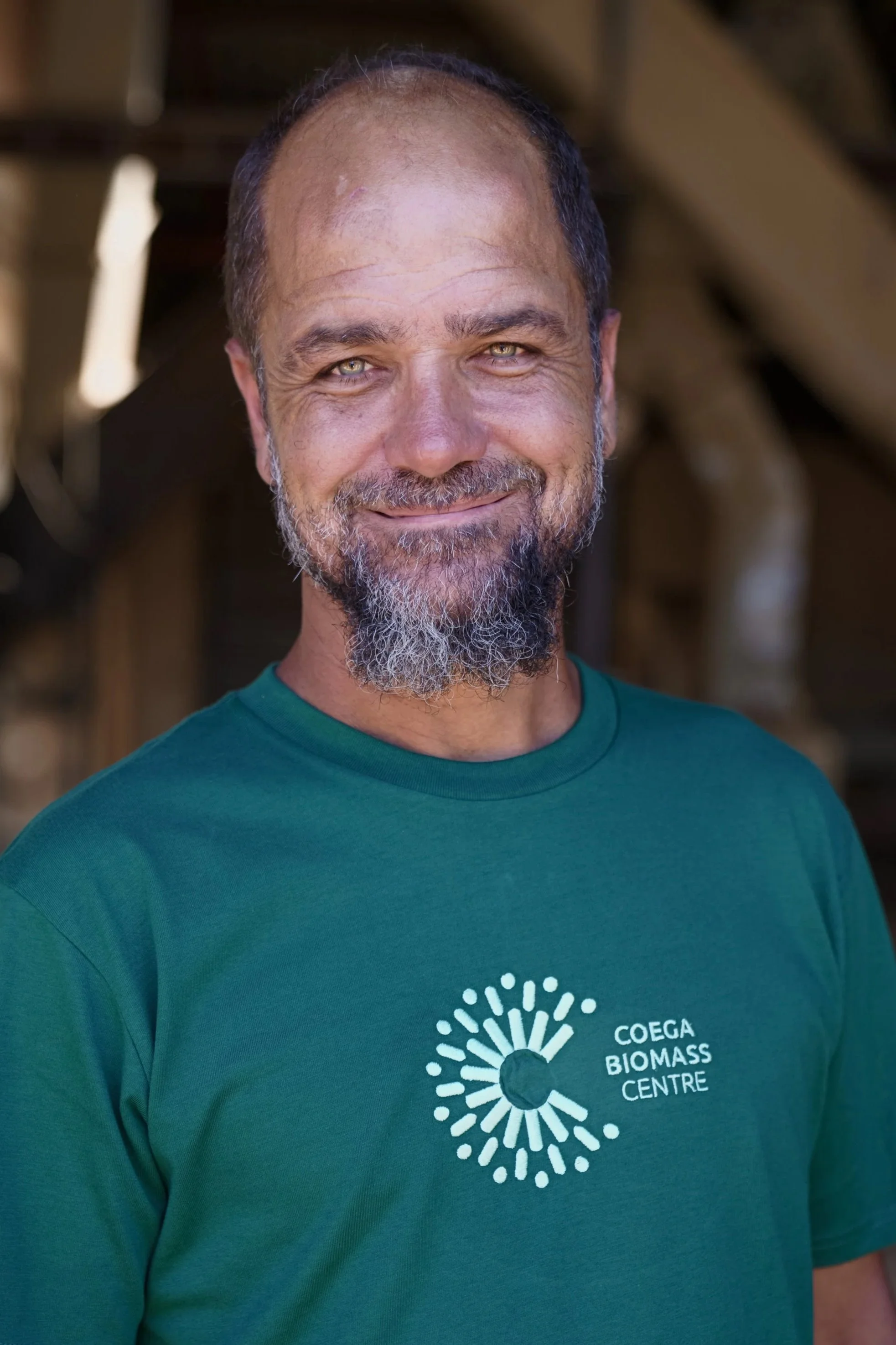 A man with a beard and short hair, smiling, wearing a teal t-shirt with the logo and text 'Coega Biomass Centre' on it, standing outdoors.