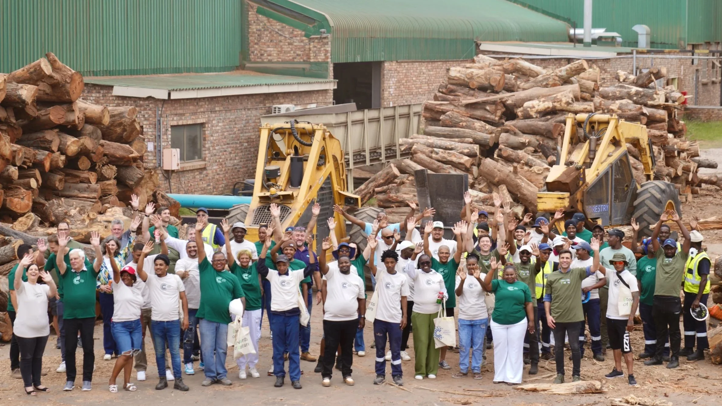 Group of people standing in front of a construction site with logs and heavy machinery, waving at the camera.