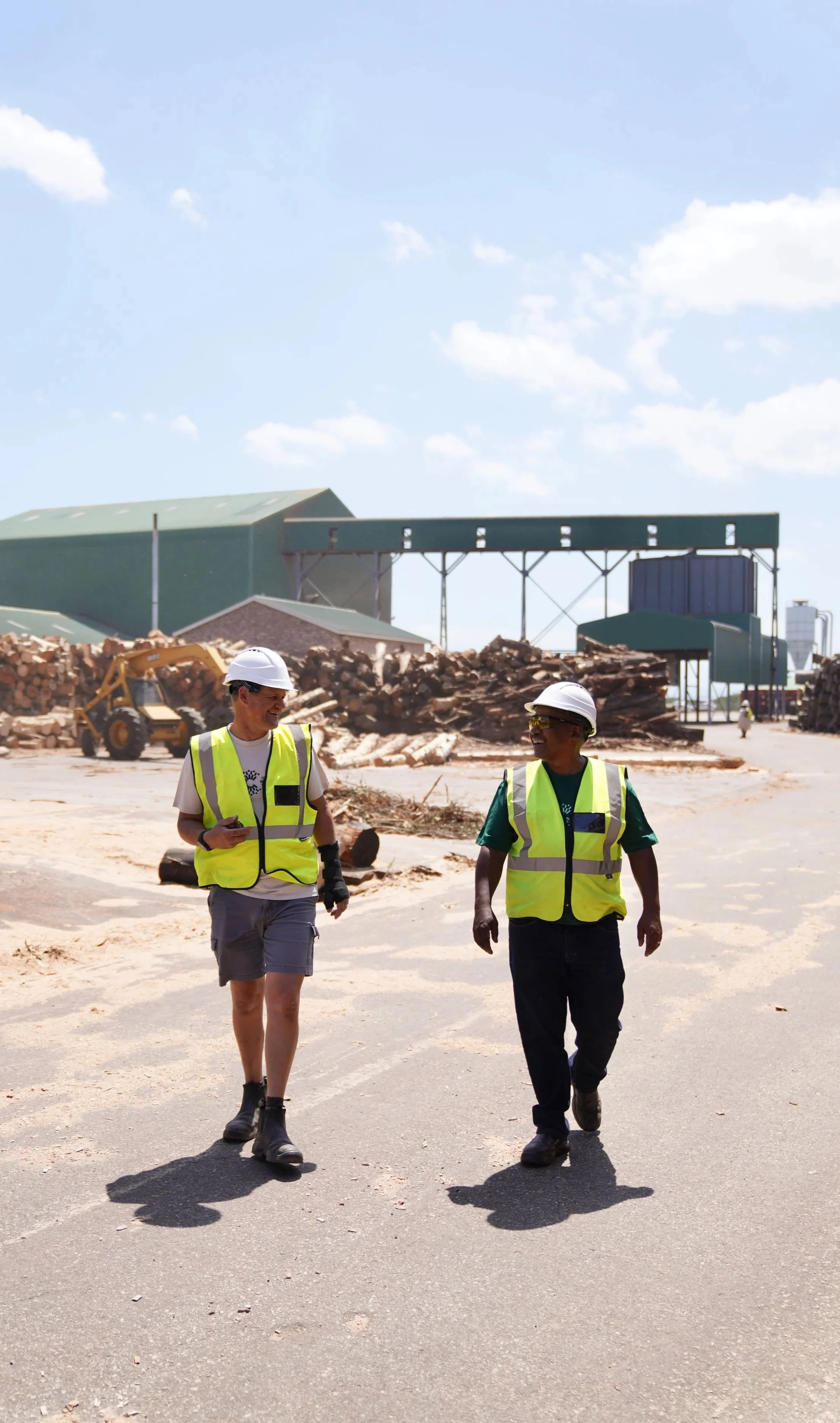 Two construction workers wearing hard hats and yellow safety vests walking and talking on a construction site with debris and machinery in the background.