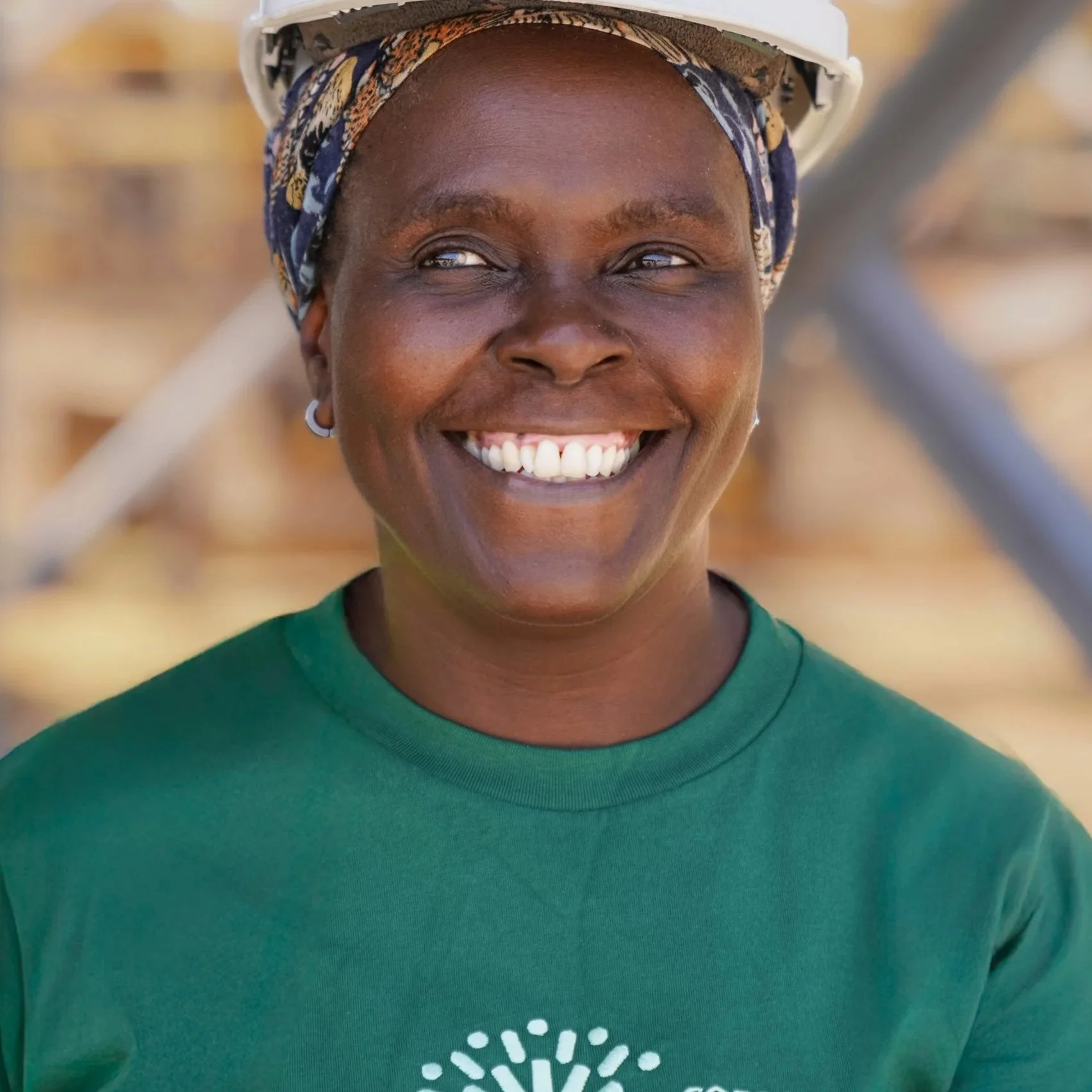 A woman smiling outdoors in front of a wooden fence, wearing a green shirt and a colorful headscarf, with a helmet on her head.