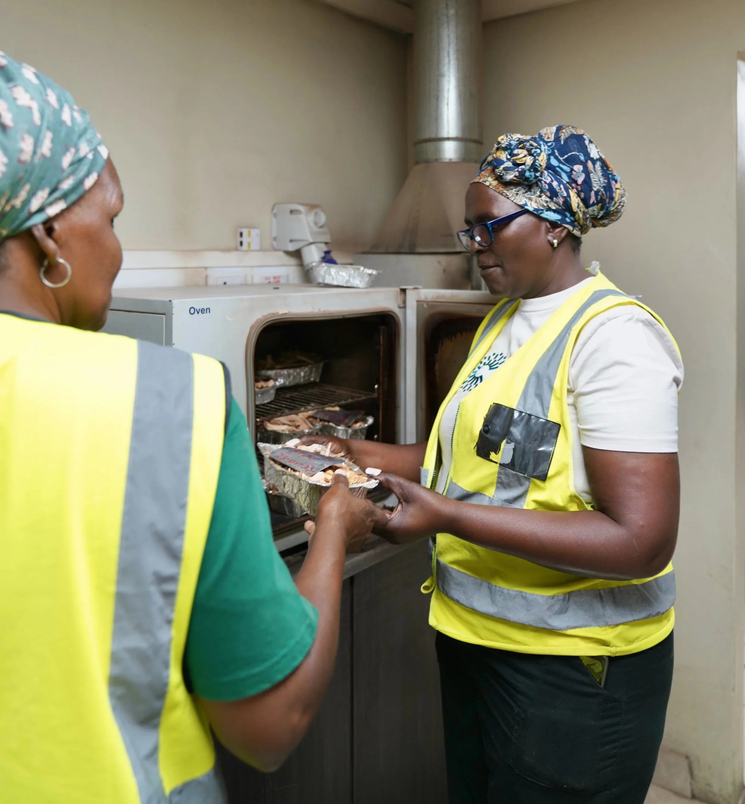 Two women wearing yellow safety vests are testing the moisture content of biomass pellets and putting the pellets into an oven.