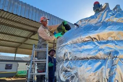 A man in a cap and gloves working on a large metallic object under a roofed outdoor structure.