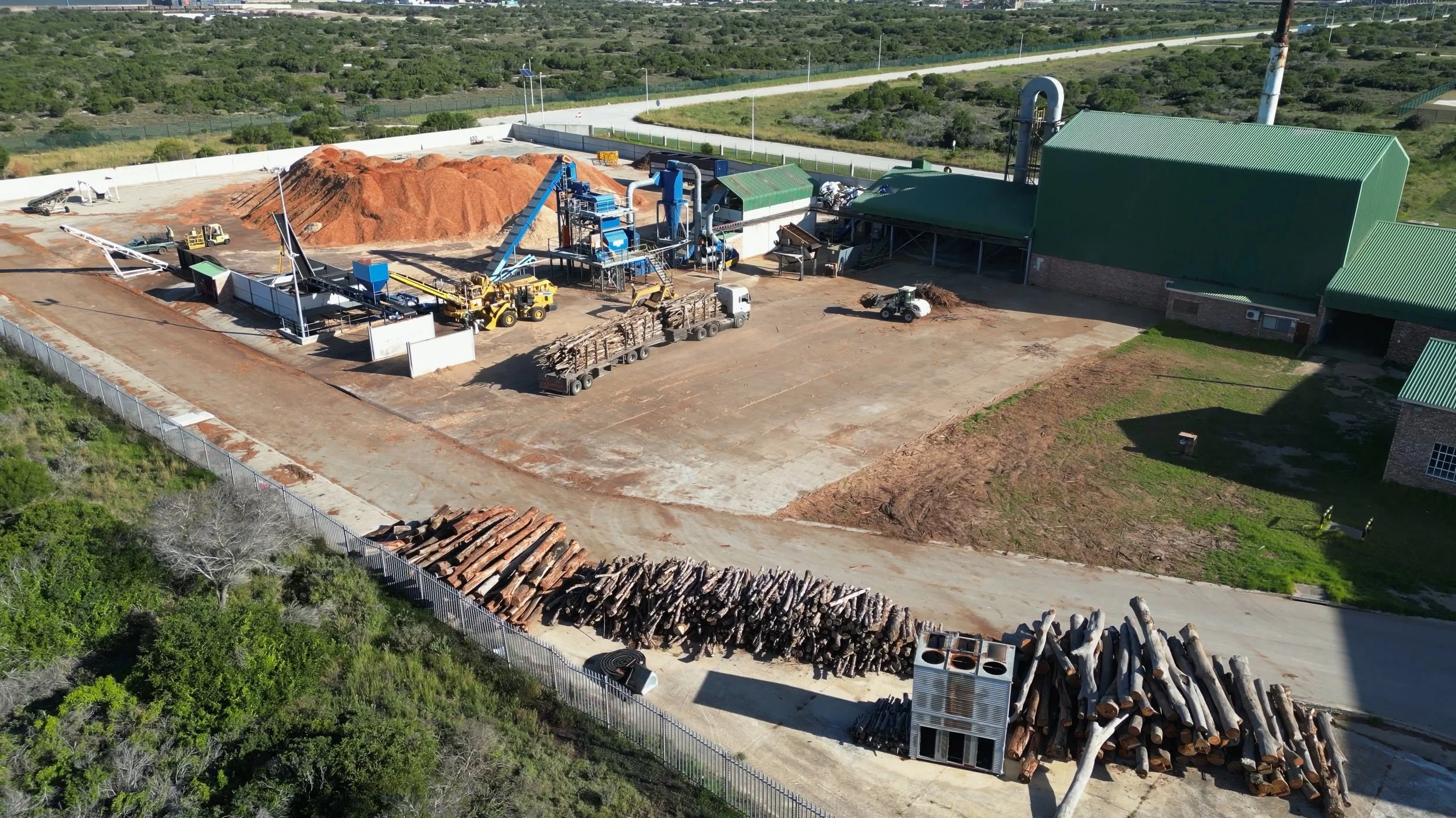 An industrial facility with machinery, trucks, and a large pile of wood chips or mulch, surrounded by greenery and a fence.