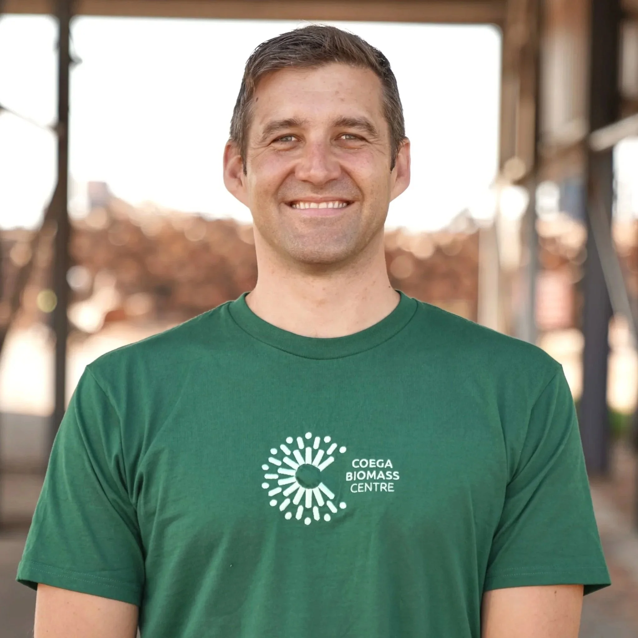 A smiling man standing outdoors, wearing a green t-shirt with a logo that reads 'Coega Biomass Centre'.