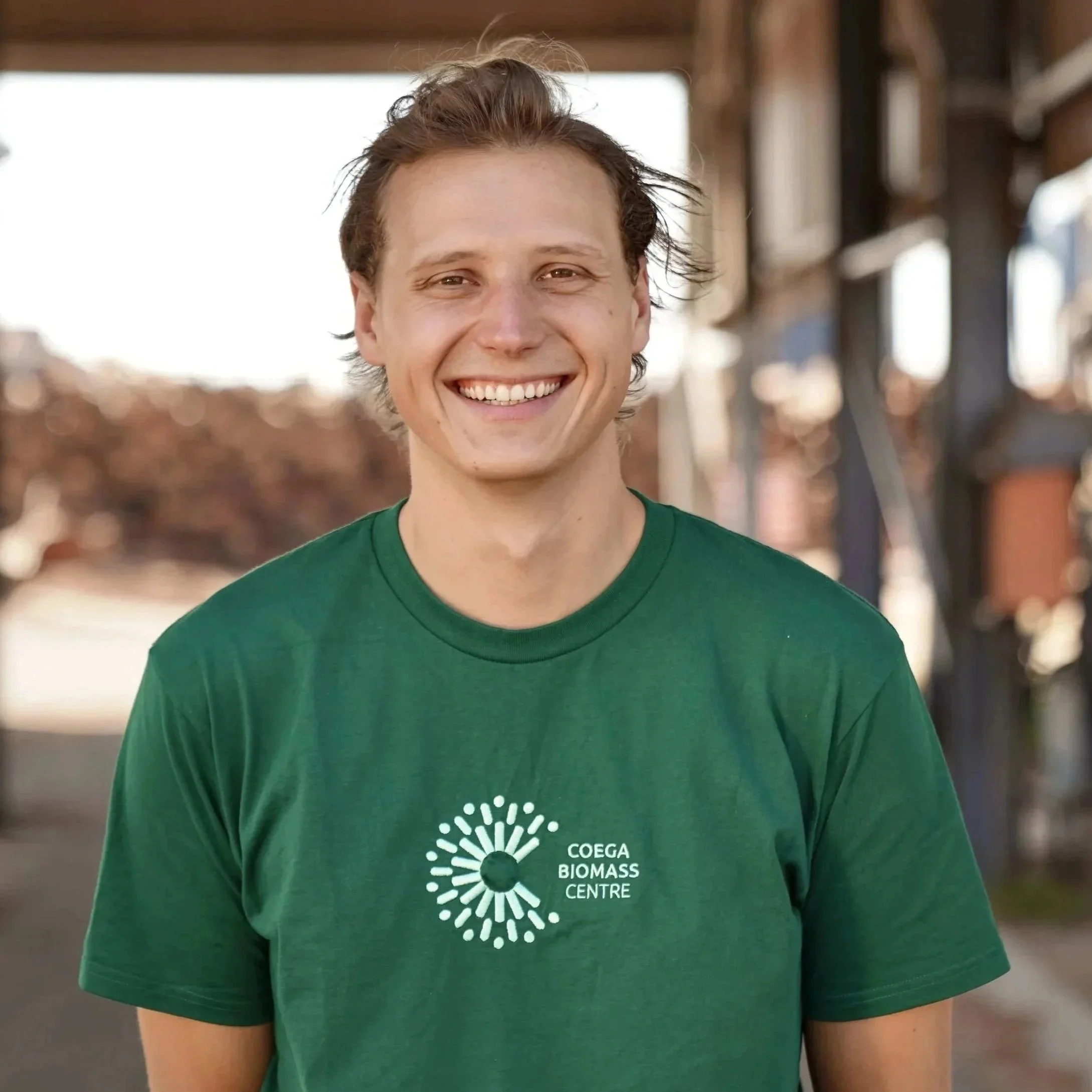 A smiling young man with light brown hair, wearing a green T-shirt with a logo that reads 'COEGA BIOMASS CENTRE,' standing outdoors in a sunny area with blurred trees and structures in the background.