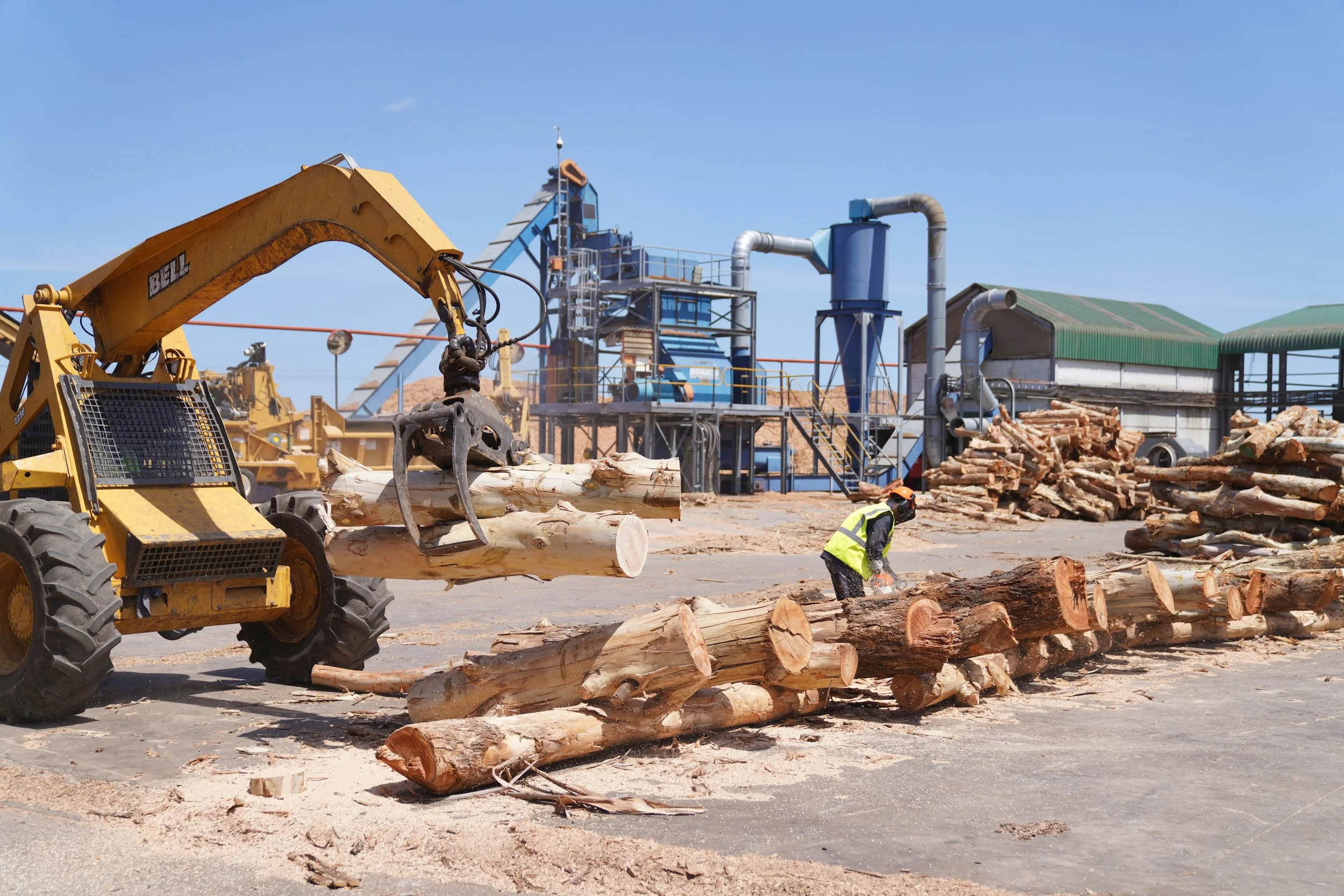 A logging yard with a large yellow forestry machine lifting logs, a worker cutting logs on the ground, and stacks of cut wood with industrial buildings and machinery in the background under a clear blue sky.