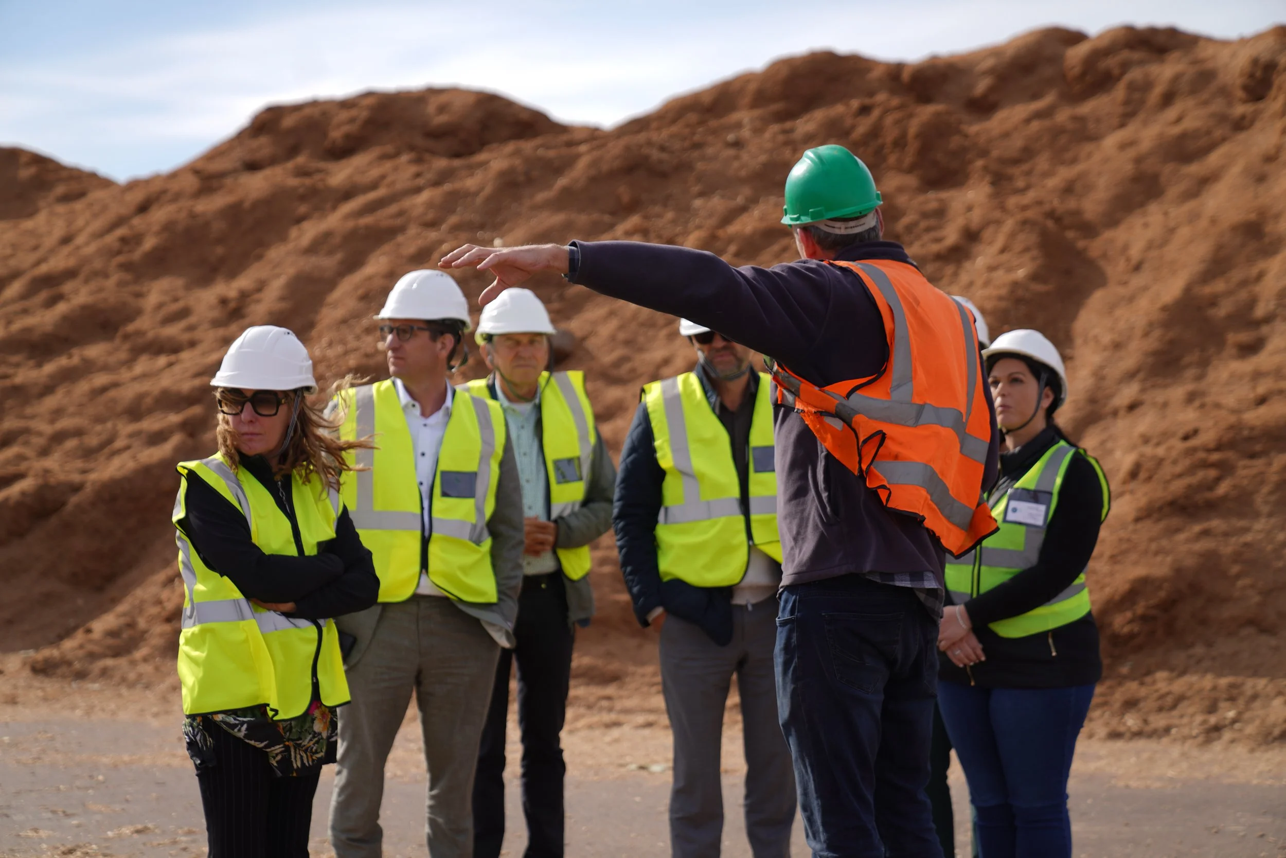 A group of construction professionals, all wearing safety helmets and yellow safety vests, listening to a man with a green helmet and orange safety vest giving a briefing outdoors near a dirt mound.