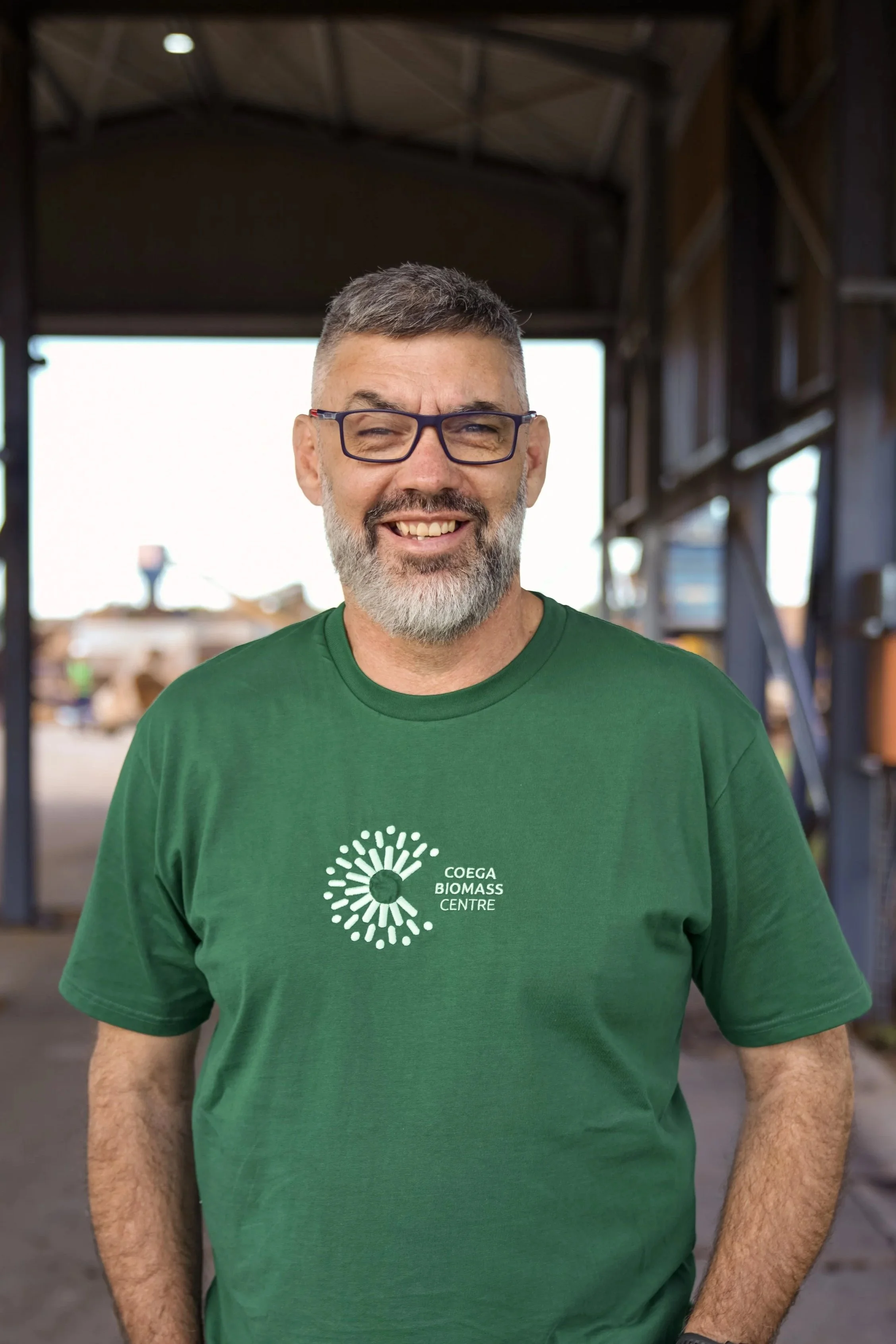 A smiling man standing outdoors, wearing a green t-shirt with a logo that reads 'Coega Biomass Centre'.