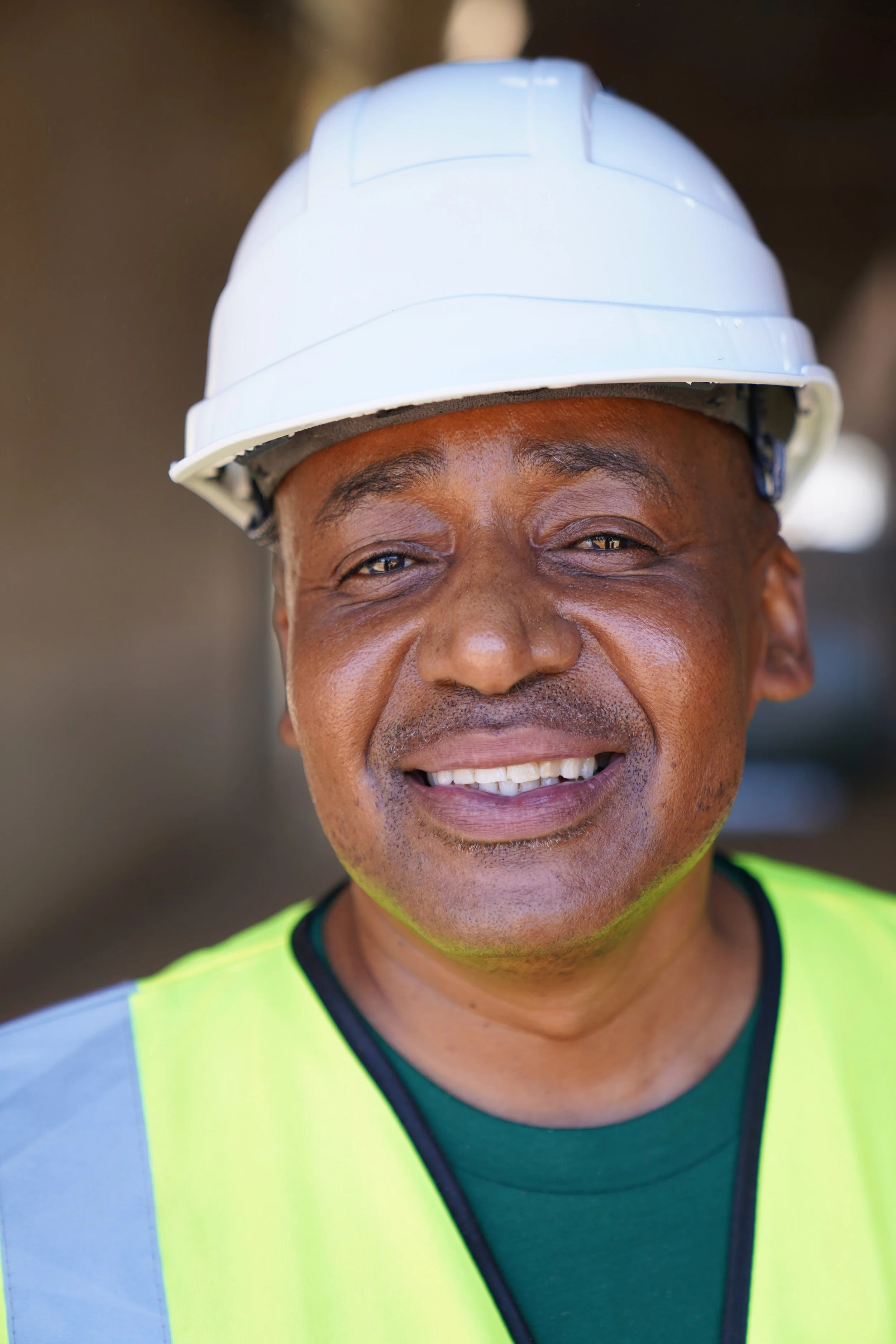 Close-up of a smiling construction worker wearing a white safety helmet and a fluorescent yellow safety vest.