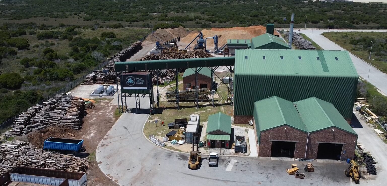 Aerial view of a biomass pellet facility with large green-roofed buildings, wood logs stacked on the sides, and industrial equipment in operation.