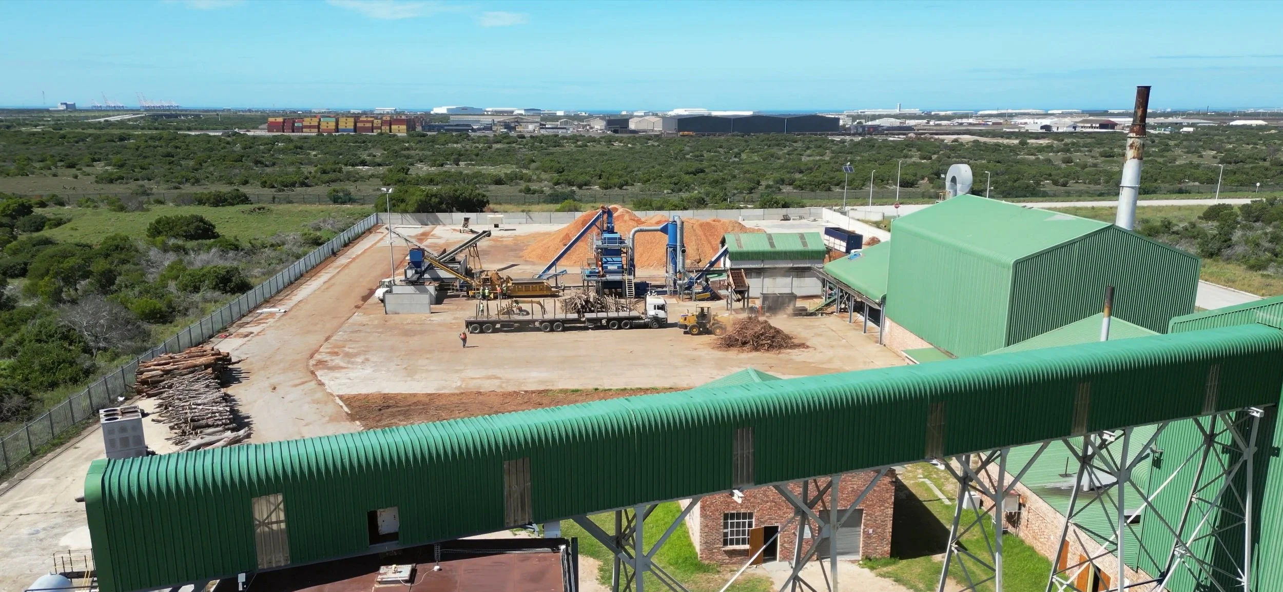 An industrial biomass pellet plant with green metal roofs, machinery, and piles of logs is set in a green, open landscape with a fence and some distant buildings on the horizon.