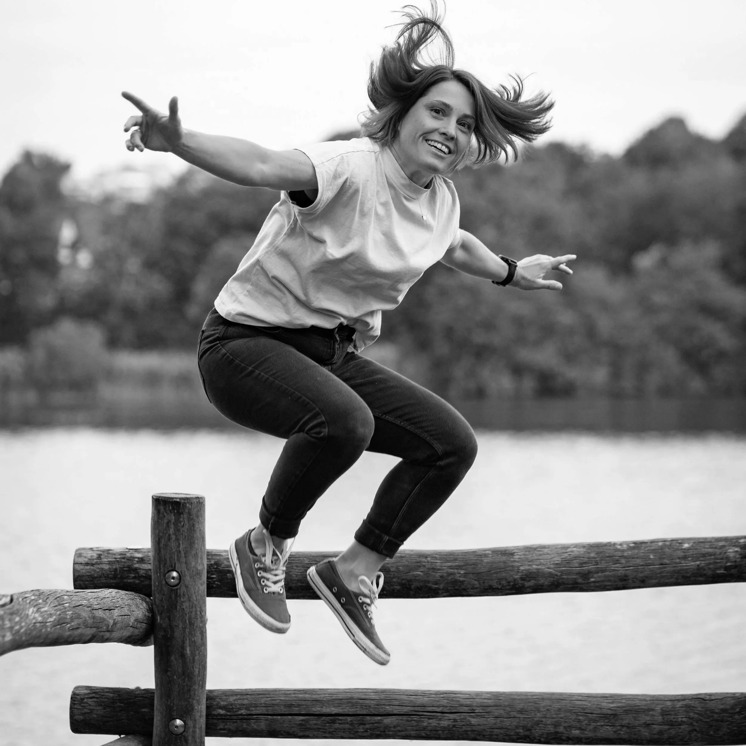 Frau springt lachend über einen Holzzaun an einem See, schwarze und weiße Fotografie.