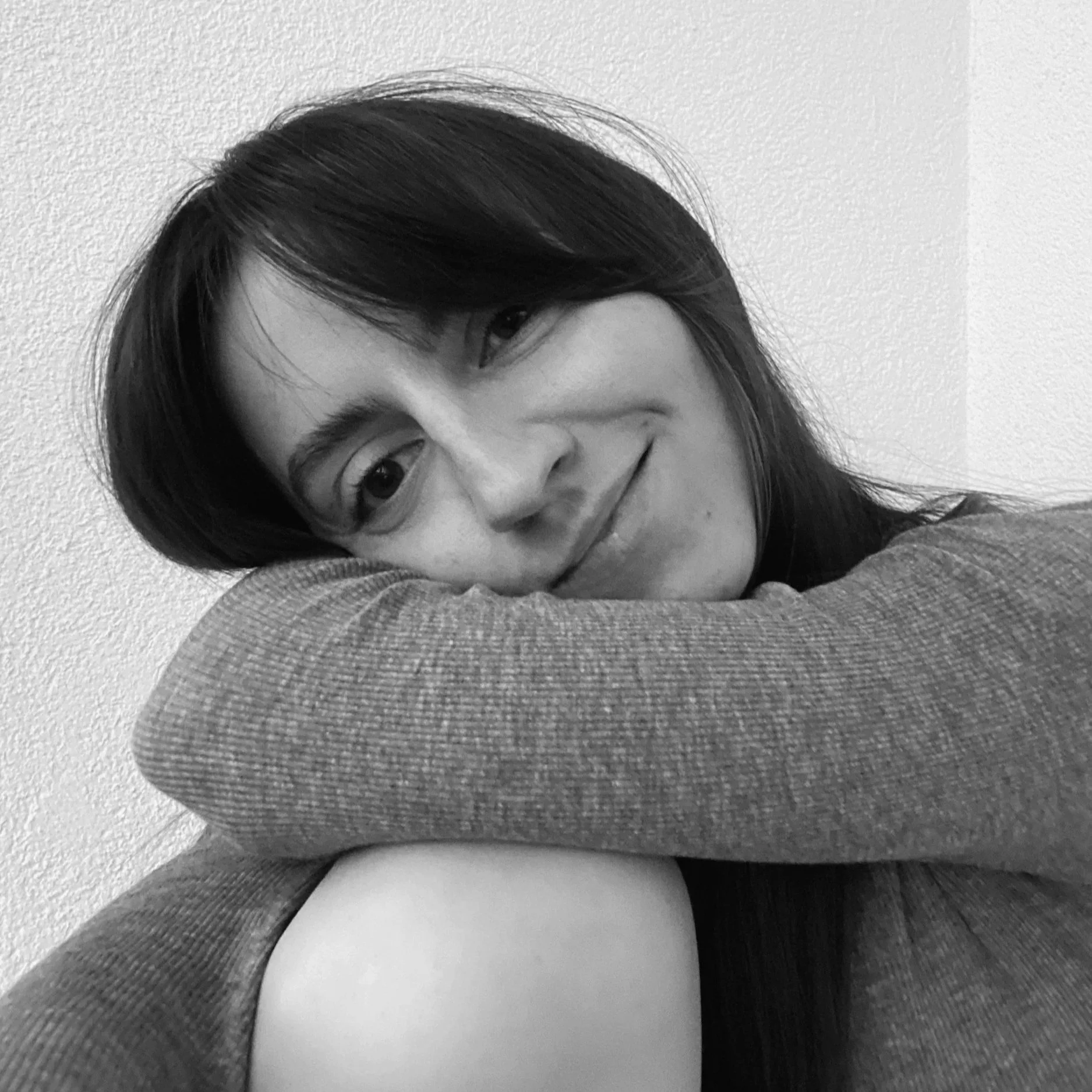 Schwarz-Weiß-Foto eines lächelnden Mädchens mit dunklen Haaren, das auf einem Arm ruht, vor einer weißen Wand.