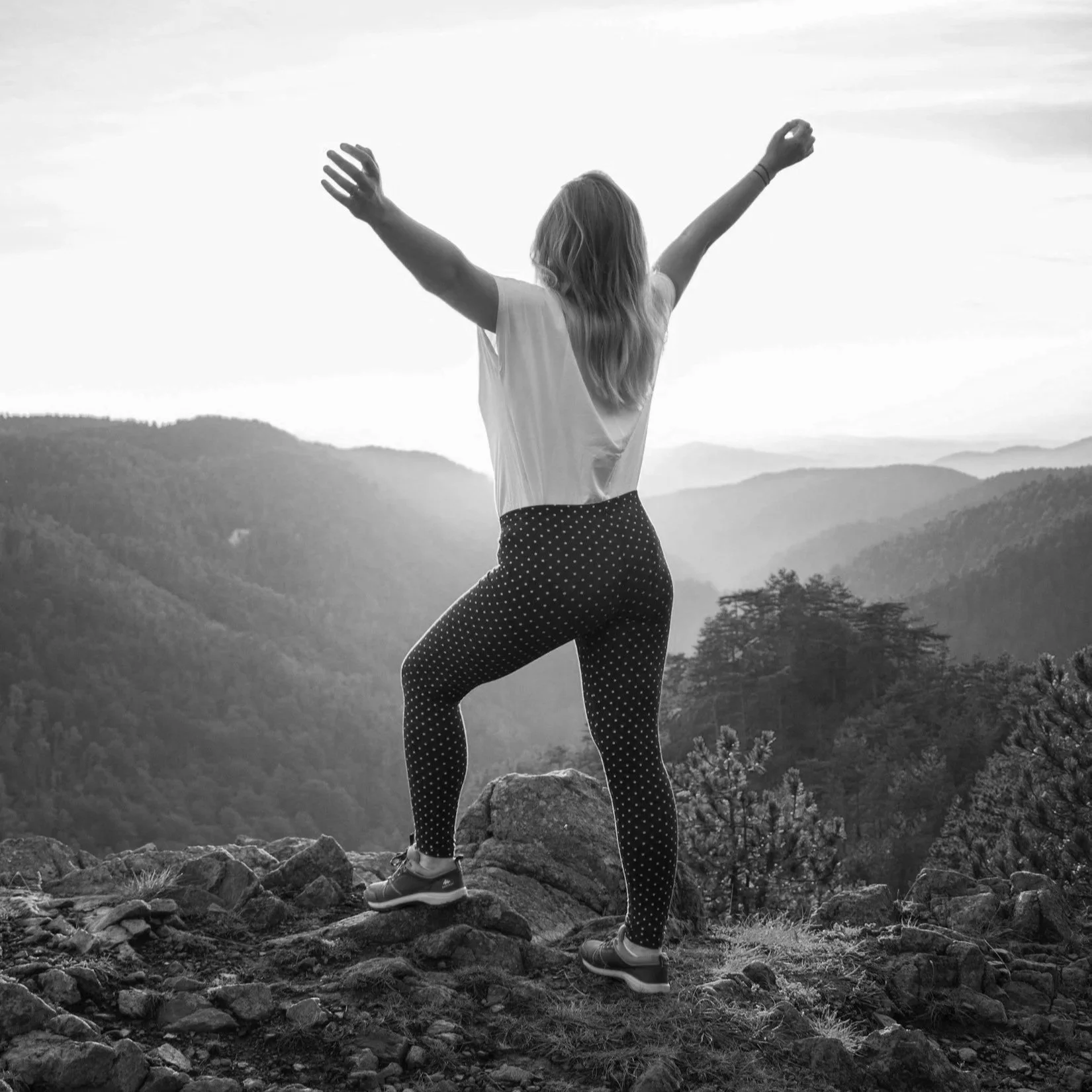 Frau auf einem Berggipfel mit erhobenen Armen, Blick auf Berge bei Sonnenaufgang, in Schwarz-Weiß.