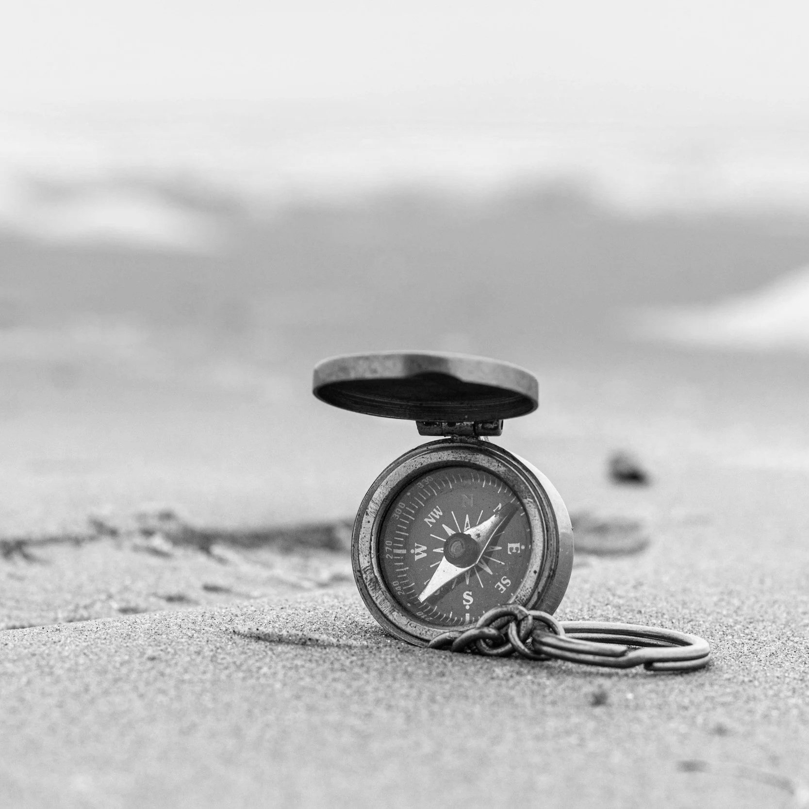 Ein Kompass liegt im Sand, mit einer Kette verbunden, am Strand bei einer Meereslandschaft, schwarz-weiß aufgenommen.