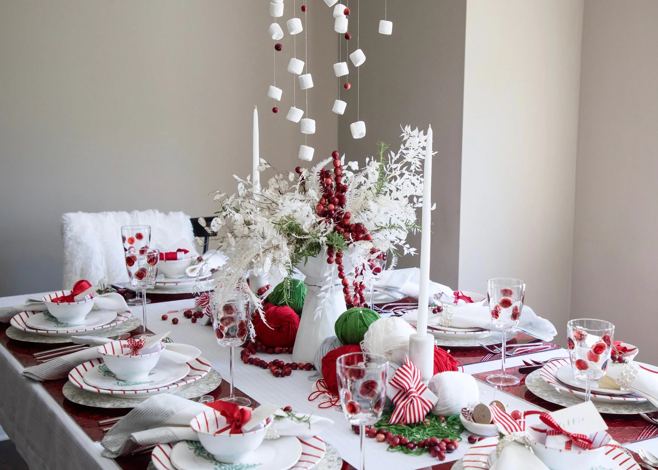 Festively decorated Christmas dining table with white, red, and green accents, featuring a floral centerpiece with white and red ornaments, candles, glassware with red berries, and coordinated tableware and napkins.