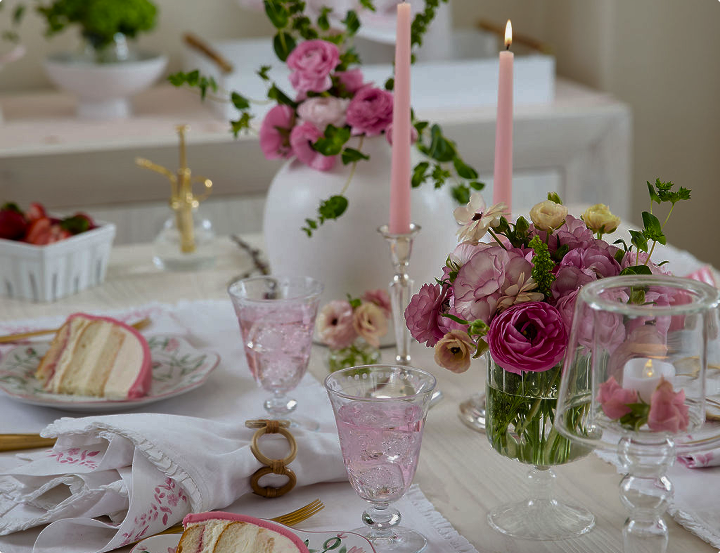A table decorated with pink flowers in vases, pink candles, pink-tinted glassware, and a slice of layered cake on a floral plate, set for a celebration or tea party.