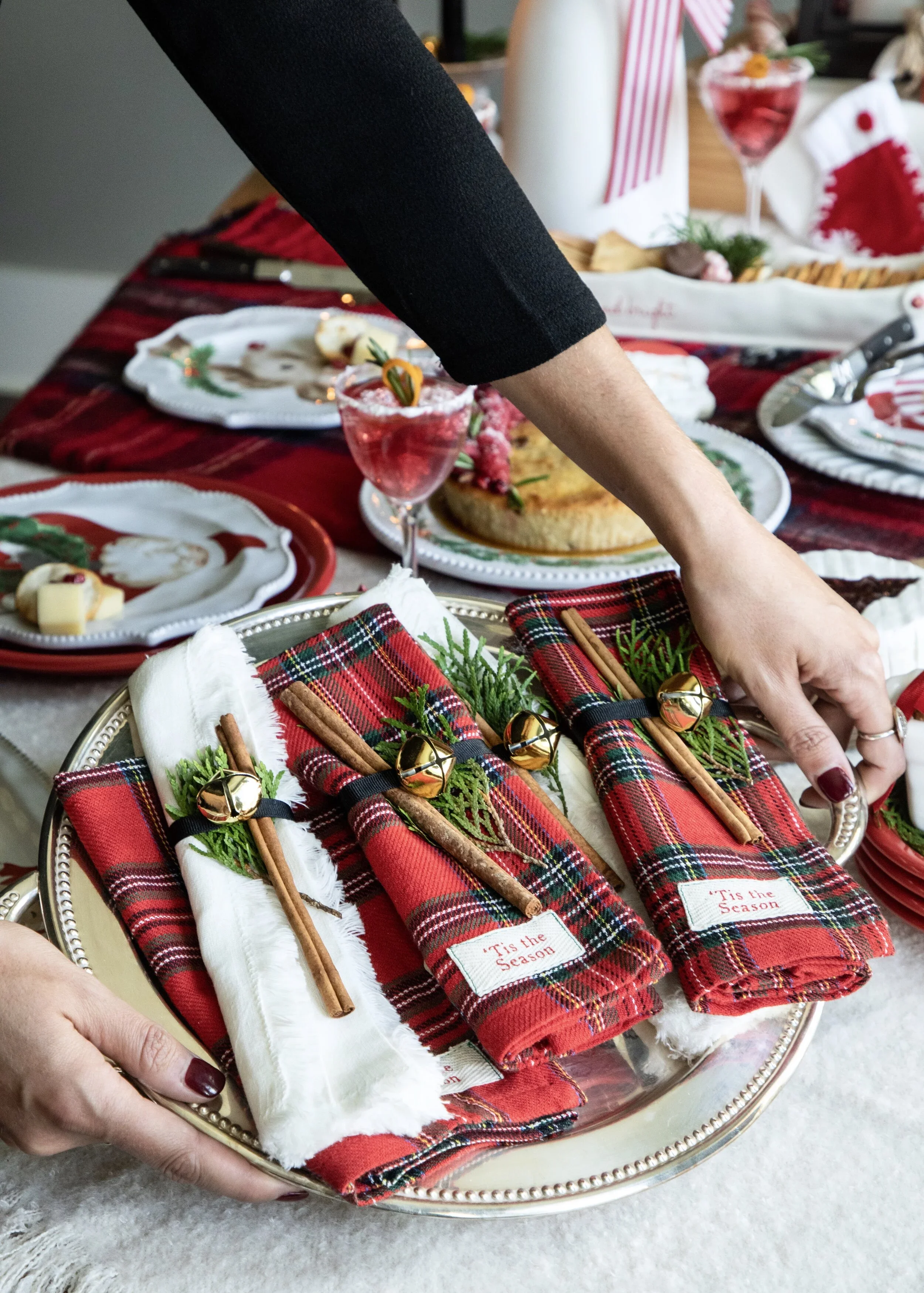 A person setting a holiday table with festive plaid napkins, cinnamon sticks, and sprigs of greenery, with a Christmas cake and holiday glasses in the background.
